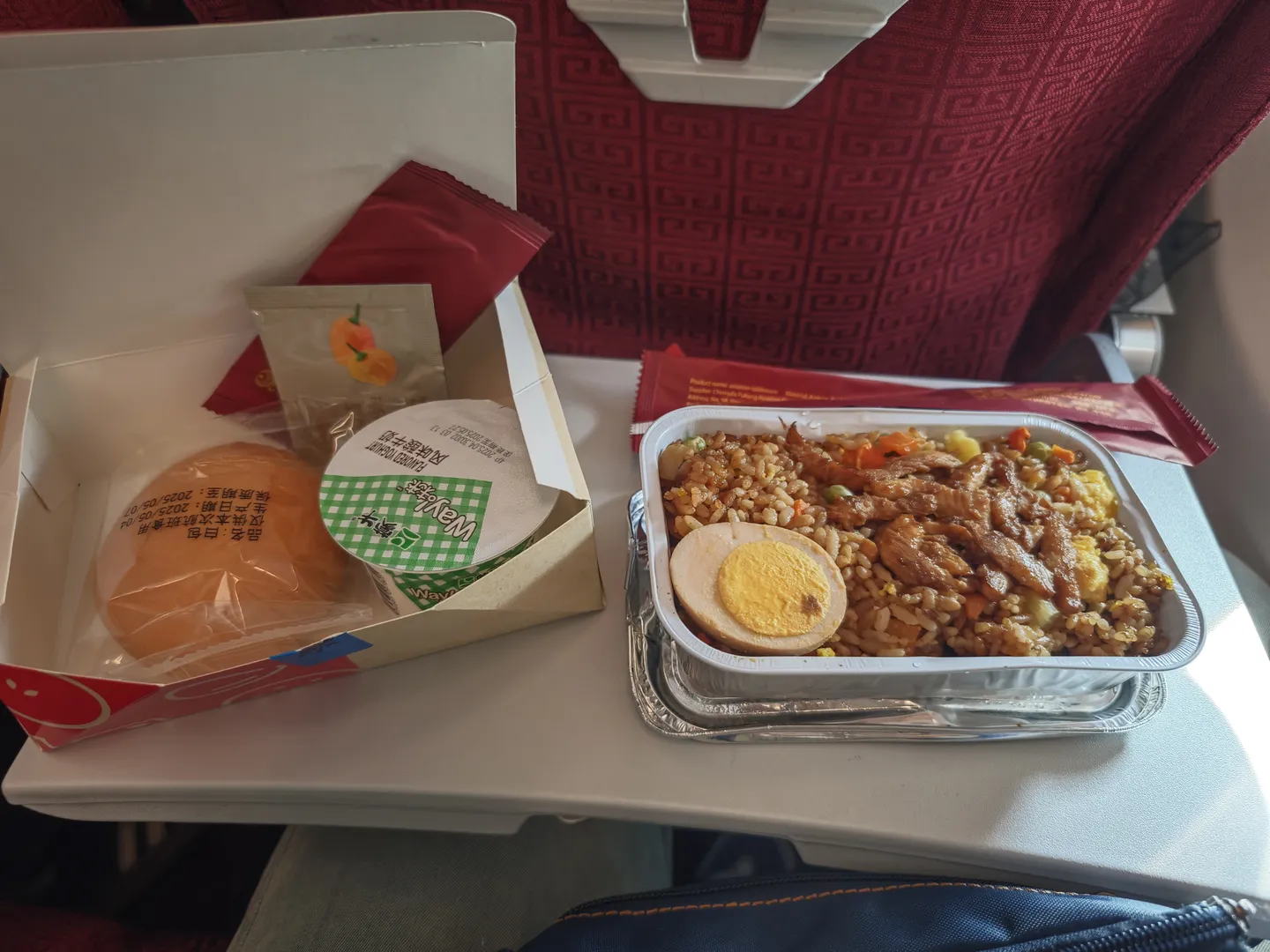 飞机餐盒放在小桌板上,包含炒饭和配菜。炒饭中有肉块、蔬菜和半个煮鸡蛋。旁边有一个包装好的面包。