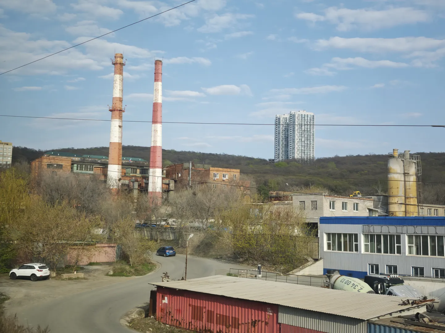 工业区景象,有两座高大的红白相间的烟囱。前景有道路和停放的车辆,背景有树木和高层建筑。工业设施和住宅楼并存。
