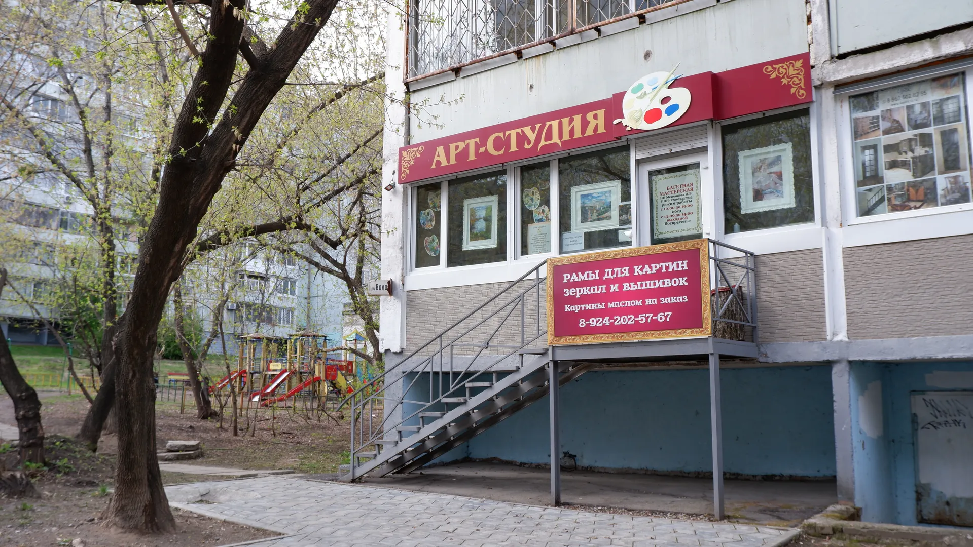 一个艺术工作室的外观,红色招牌上写着“АРТ-СТУДИЯ”,门口有楼梯和展示的画作。旁边是树木和一个儿童游乐场,游乐场设备色彩鲜艳。建筑物外墙为粉色和白色,底层有商店招牌,前面停着几辆车。