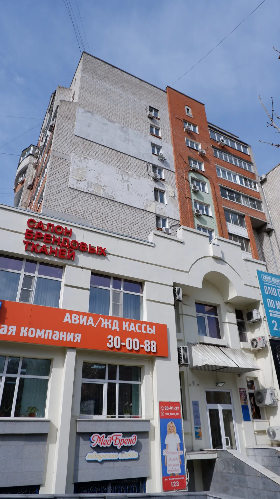 一栋高层建筑,底层有商铺招牌,上面写着“САЛОН БРЕНДОВЫХ ТКАНЕЙ”。建筑外墙部分区域剥落,窗户排列整齐。天空晴朗,电线横跨画面。