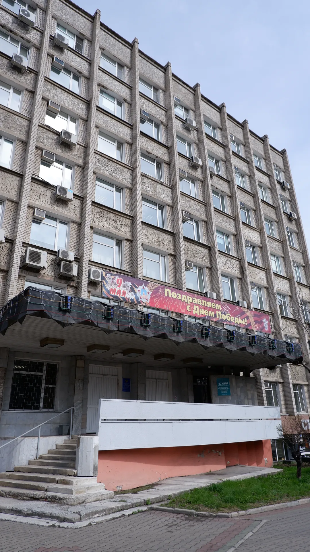 一栋多层建筑,底层有入口台阶和红色装饰。建筑正面悬挂着庆祝胜利日的横幅,窗户排列整齐。天空阴沉,地面有绿化带。