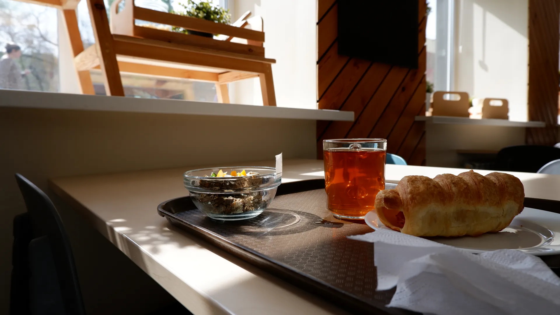 桌子上放着一个托盘,托盘上有茶杯、羊角面包和一碗食物。阳光透过窗户洒在桌子上,背景中有木质椅子和墙壁。