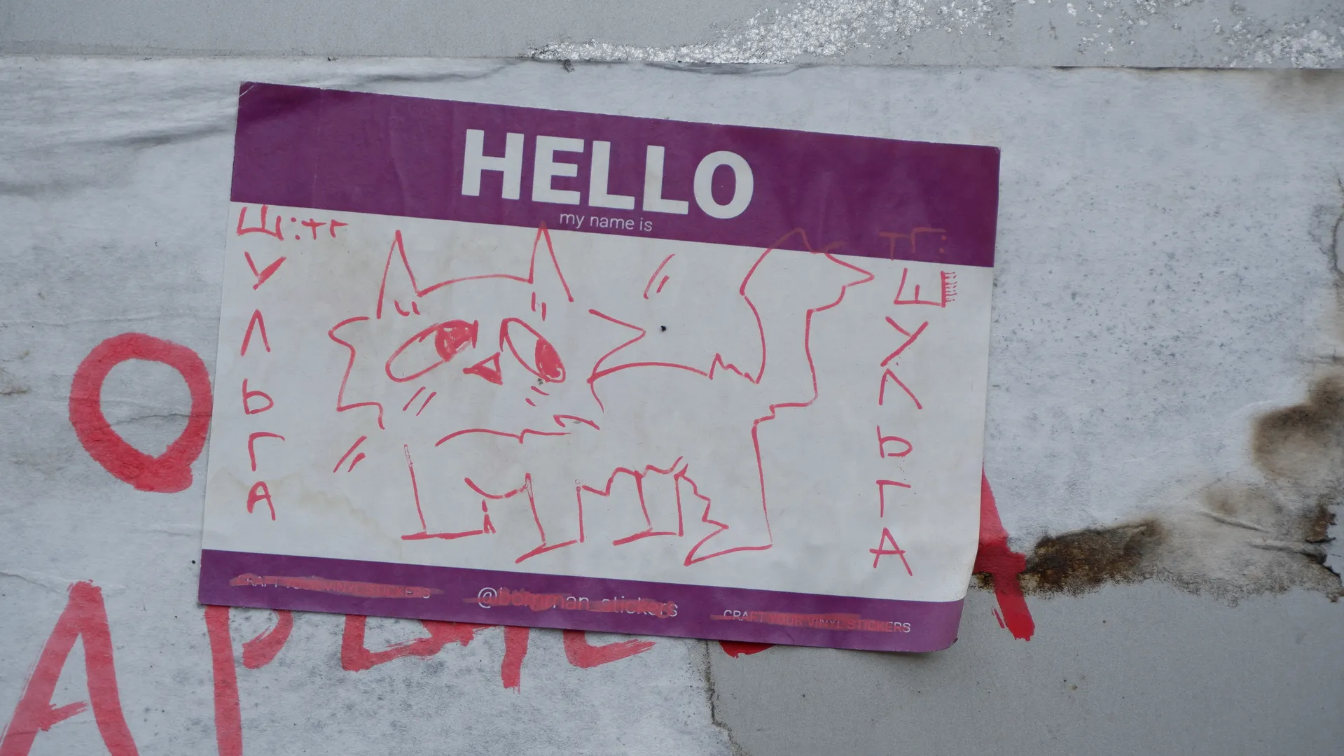 墙上贴着一张纸,纸上画着一只红色线条的猫,写着“HELLO my name is”。纸张四周有红色涂鸦,背景是灰色墙面。