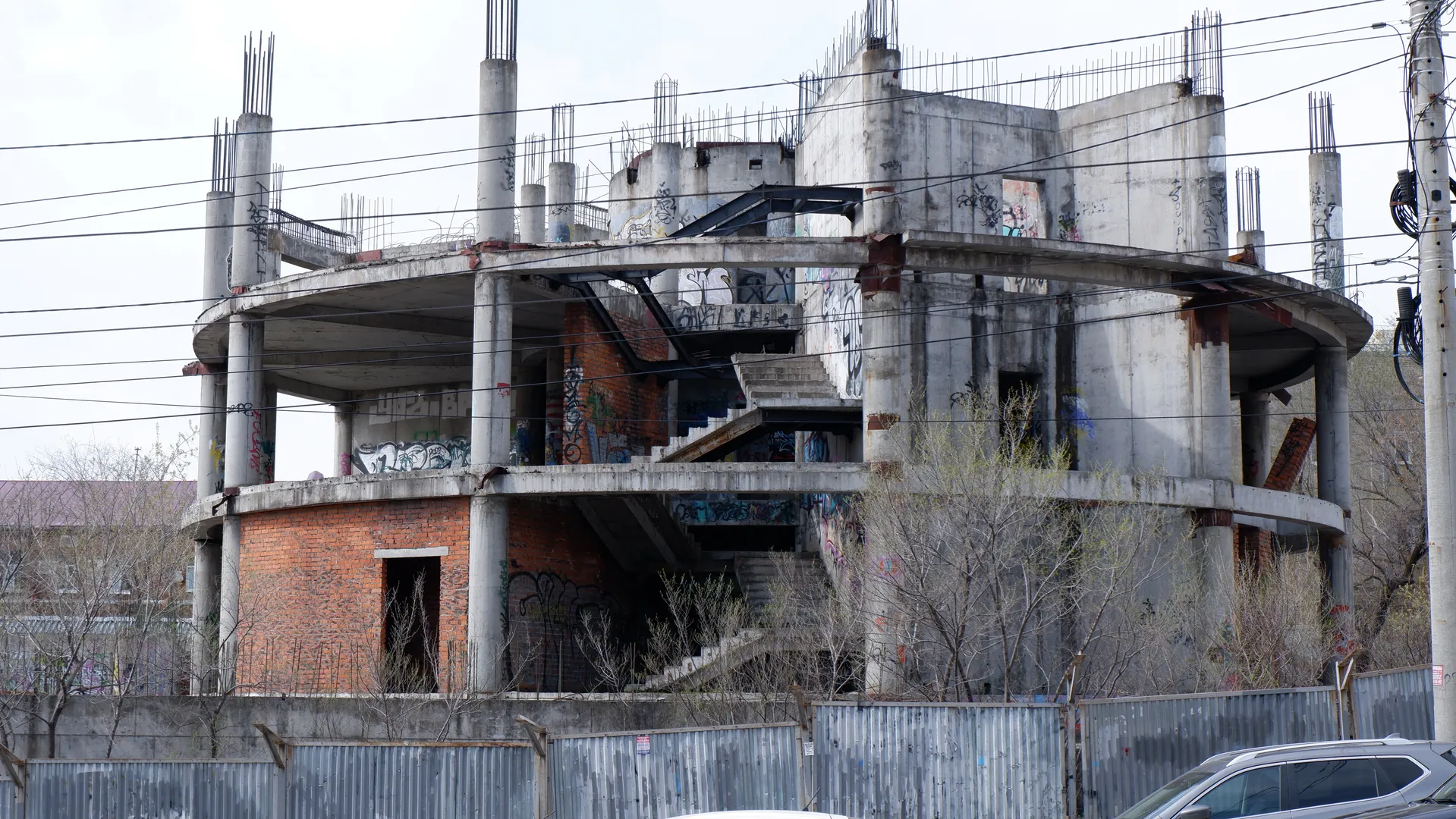 一栋未完工的建筑,结构裸露,墙上有涂鸦。建筑前方有围栏,周围有树木。天空阴沉。