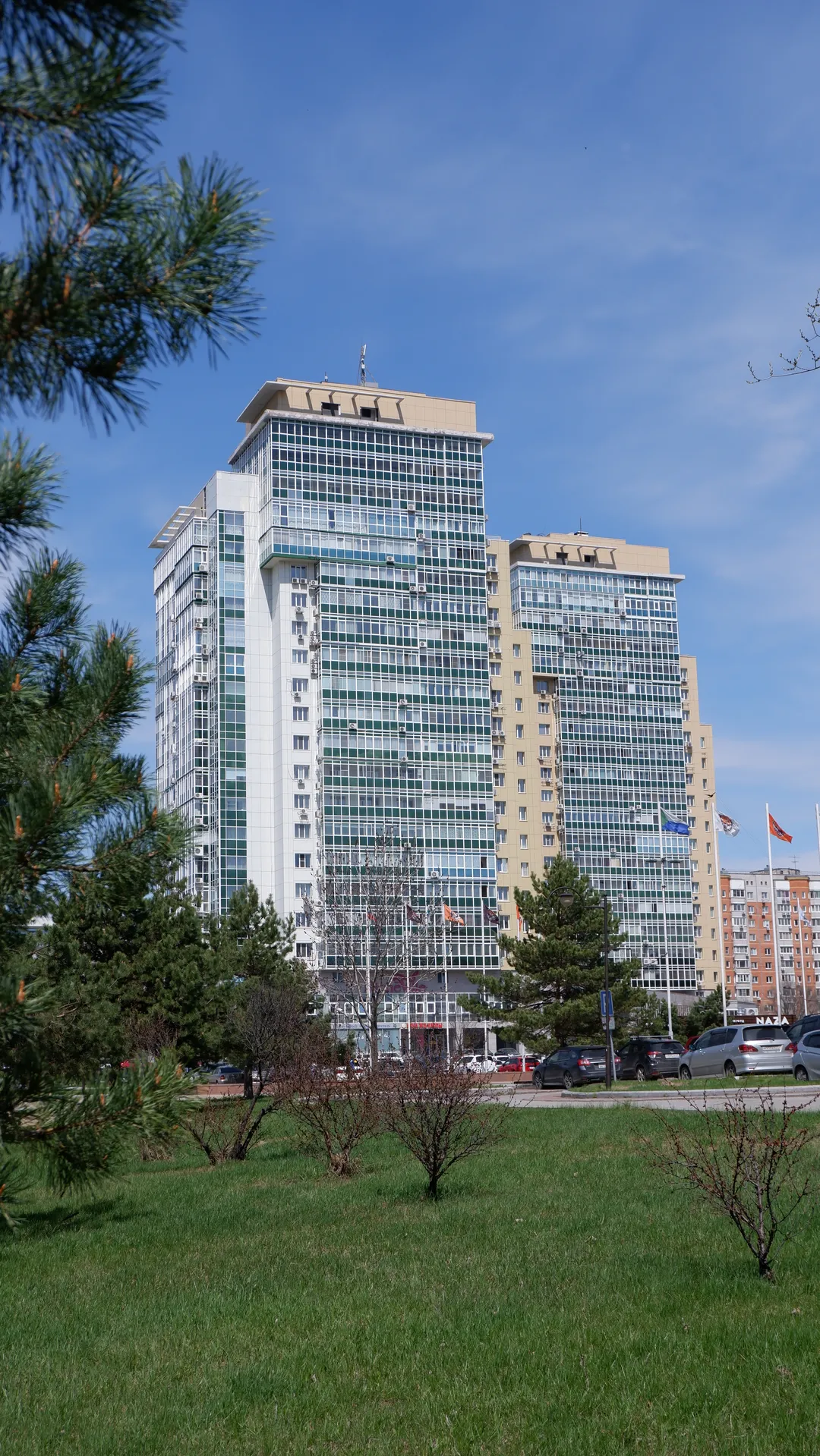 几栋高层建筑耸立在蓝天之下,建筑外墙主要是玻璃和混凝土结构。前景中有树木和草地,建筑底部有一些车辆停放。天空晴朗,没有云朵。