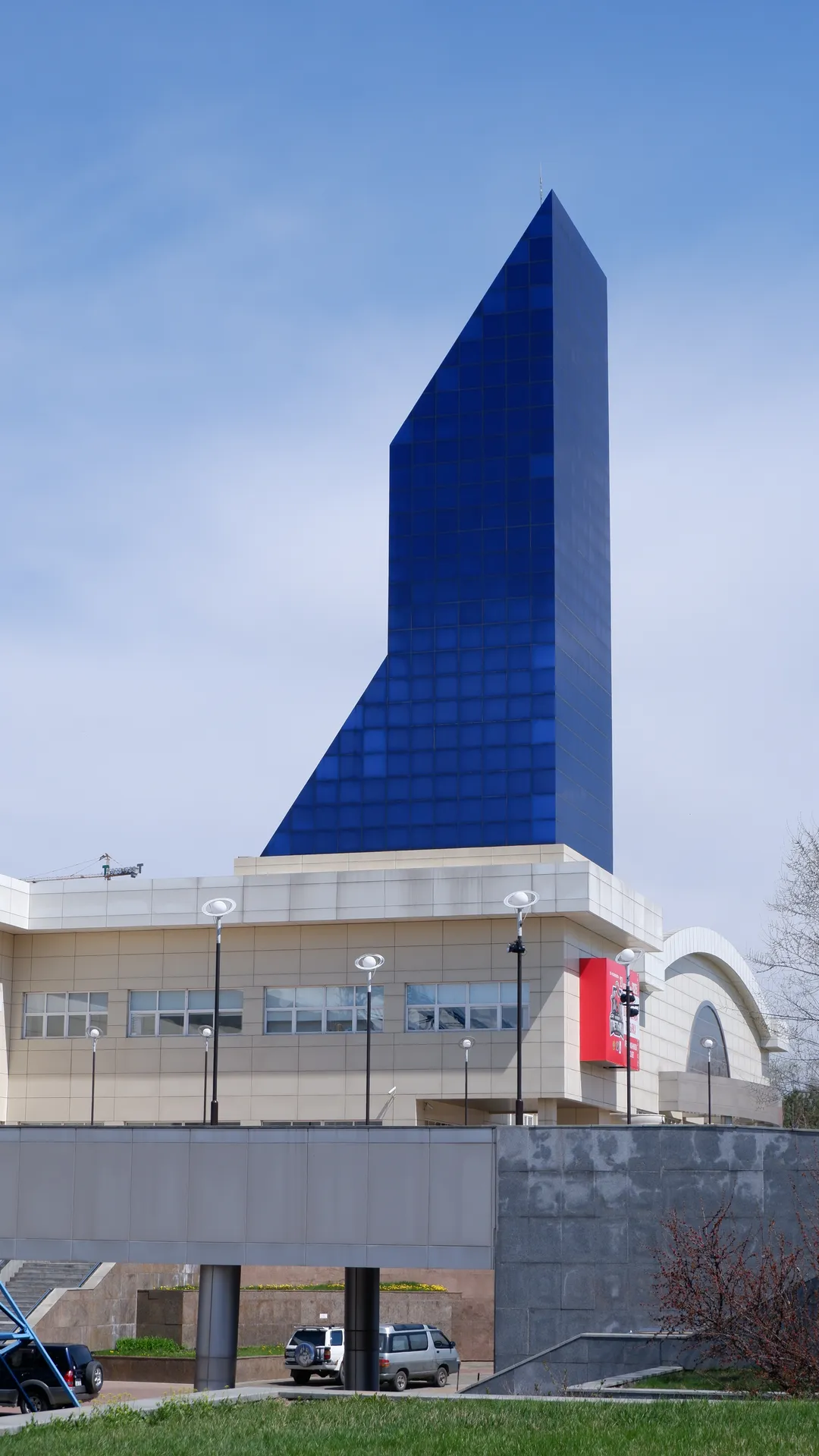 一座现代建筑有一个蓝色玻璃金字塔状的塔楼。建筑底部是浅色石材结构,前面有几盏路灯和一个广告牌。天空晴朗,背景中有一些树木和灌木。