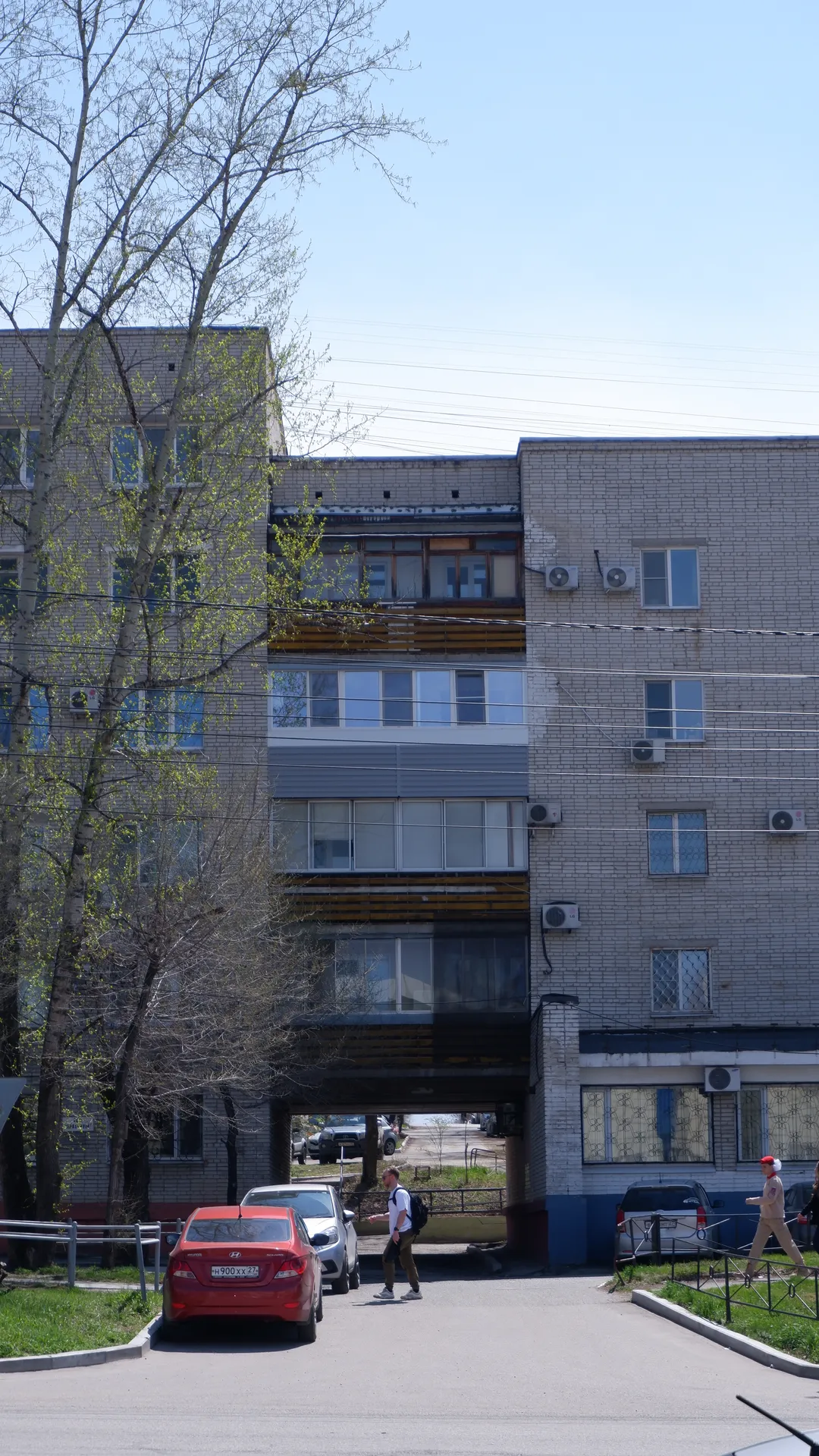 一栋多层住宅楼,外墙由砖块和混凝土构成,窗户排列整齐。楼前有一个停车场,停着几辆汽车。几个人在楼前行走,背景中有电线和树木。