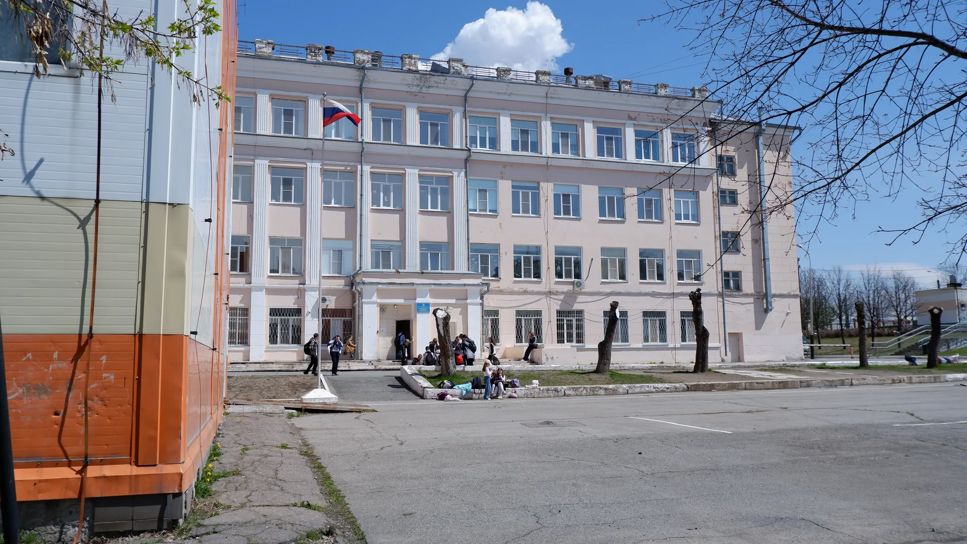 一栋多层建筑前有一个广场,建筑上飘扬着一面旗帜。几个人在建筑前活动,旁边有一棵无叶的树。天空晴朗,呈现出蓝色。
