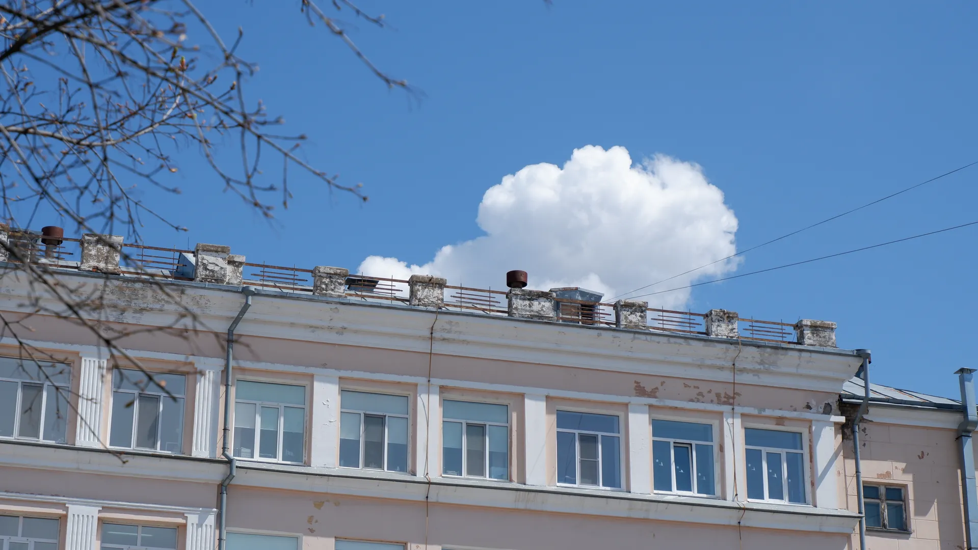 一栋多层建筑的顶部有一些通风管道和护栏。建筑外墙有多个窗户,颜色为浅色。天空中有几朵白云,树枝部分遮挡了画面。