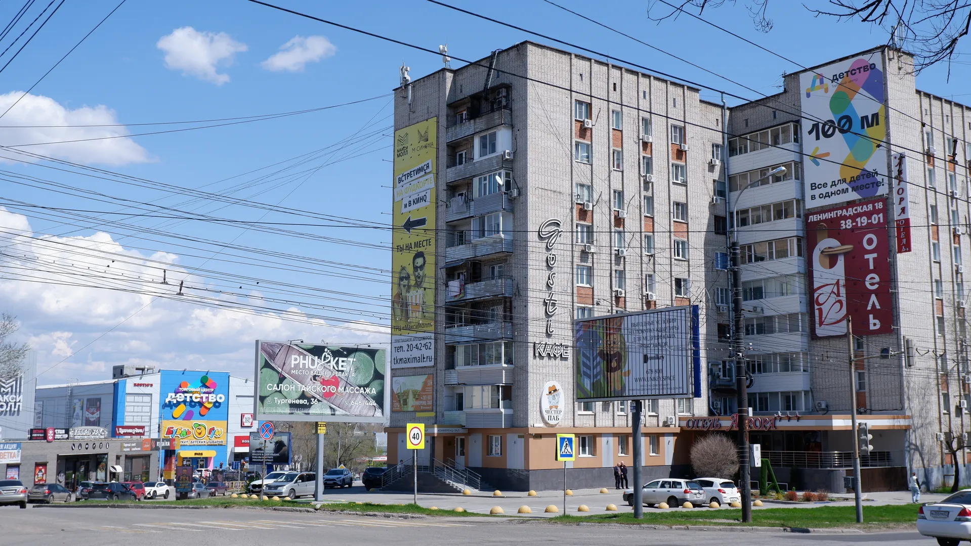 一条街道旁有多层建筑,建筑上有广告牌和招牌。街道上有行驶的车辆和交通标志。天空中有几朵白云,电线纵横交错。