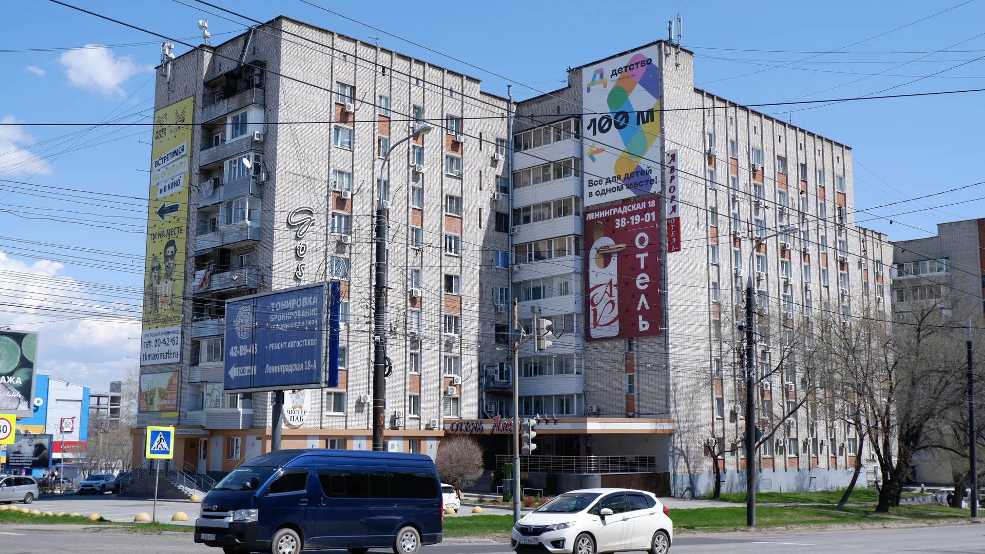 多层建筑前的街道上有行驶的车辆,建筑上有大型广告牌。广告牌内容涉及商业信息,字体醒目。天空晴朗,电线贯穿画面。