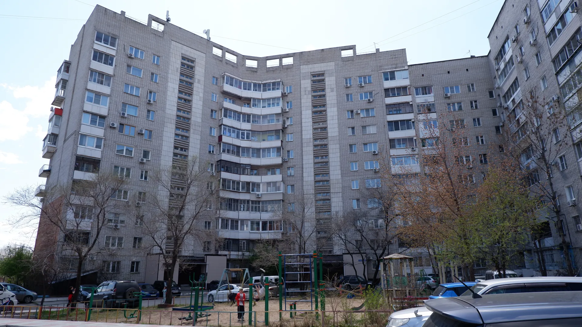一栋高层建筑,外墙为灰色瓷砖,有许多窗户和阳台。建筑前方有一个儿童游乐场,里面有滑梯和秋千。树木环绕在建筑周围。