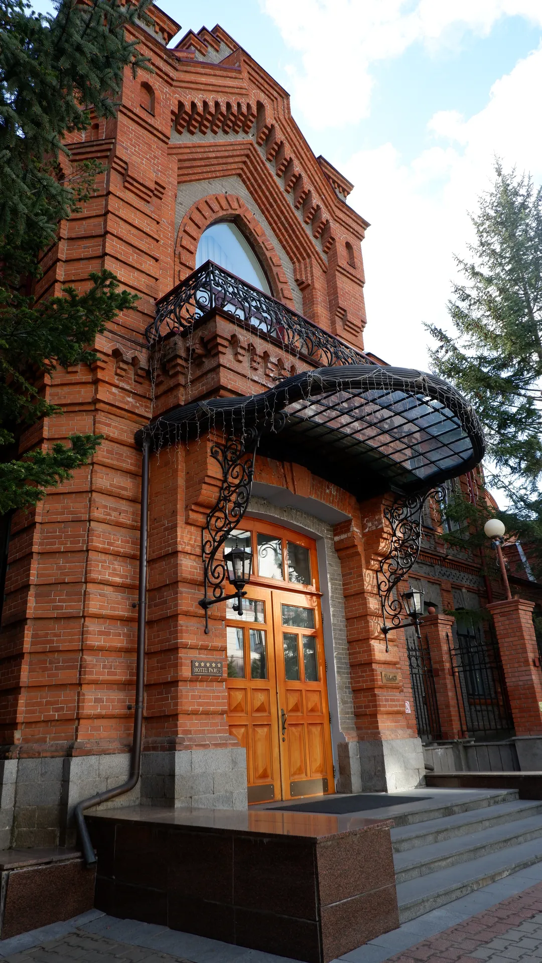 一座红砖建筑,门口有橙色木门和黑色铁艺装饰。建筑正面有拱形窗户和阳台。门前有台阶和路灯。