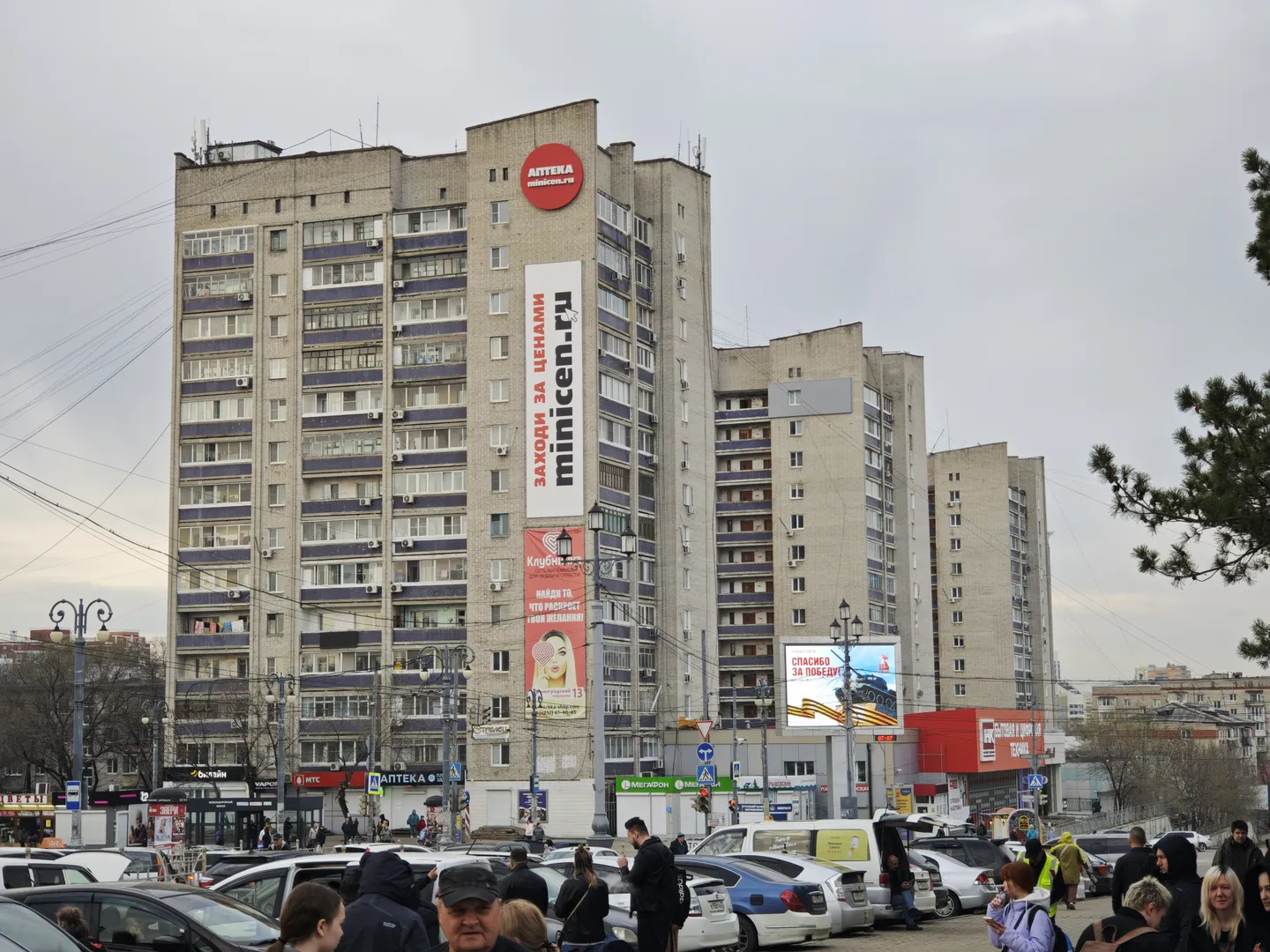 一幢高层建筑外观陈旧,外墙为灰色,窗户排列整齐。建筑上有多块广告牌,前景中停满了车辆。许多人行走在建筑前的街道上。