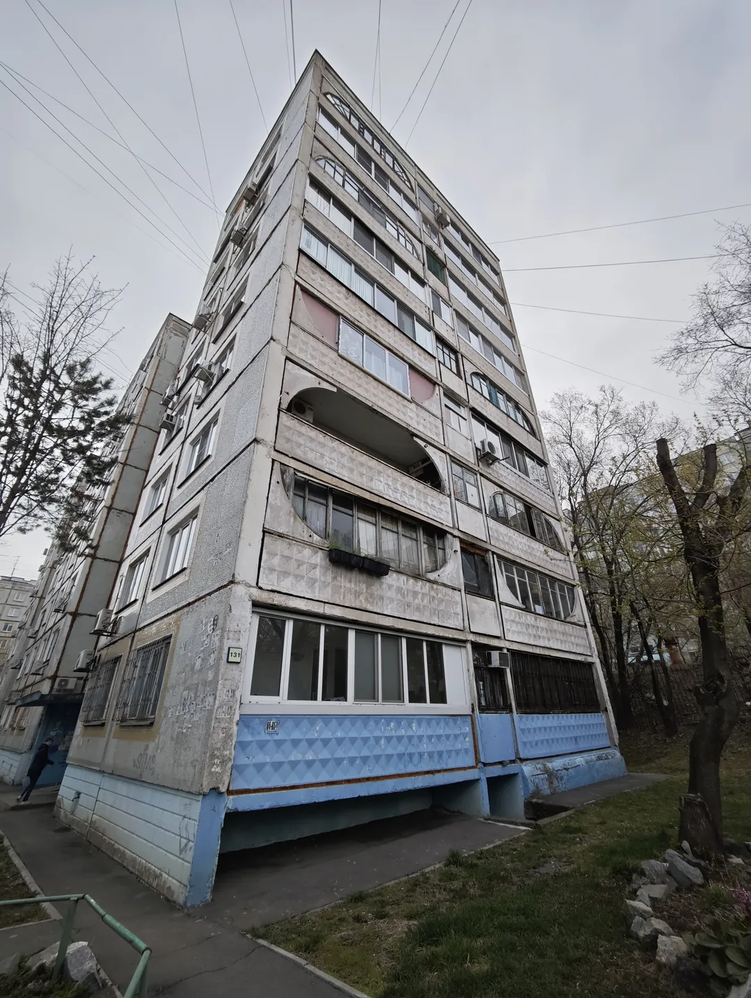 一栋高层建筑,外墙为浅色,底部有蓝色装饰。建筑有多层阳台和窗户,外墙有些许污渍。周围有树木和电线,地面有小径和草地。