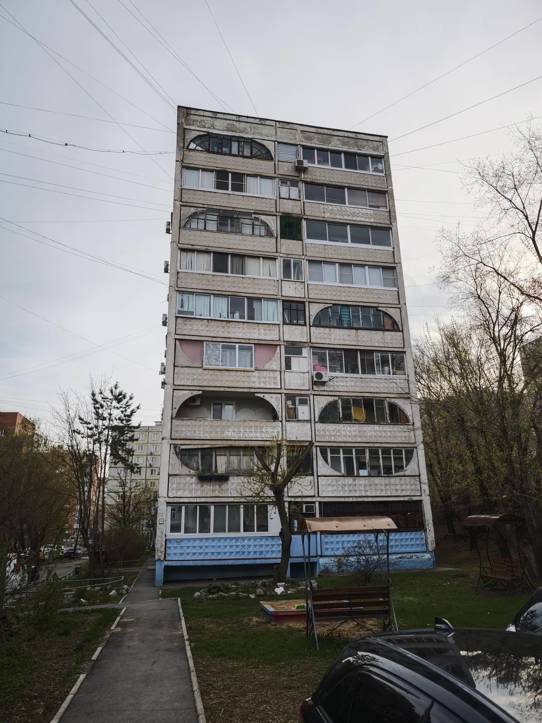 一栋高层建筑,外墙为浅色,每层都有弧形阳台。建筑底部有蓝色装饰,周围有树木和绿地。前方有一条小径,旁边停放着一辆车。