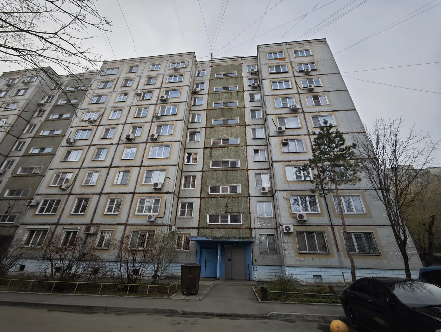一栋多层建筑,外墙为浅灰色,窗户排列整齐。建筑入口处有蓝色门,门前有垃圾桶和树木。旁边停着一辆黑色汽车,地面为水泥路。