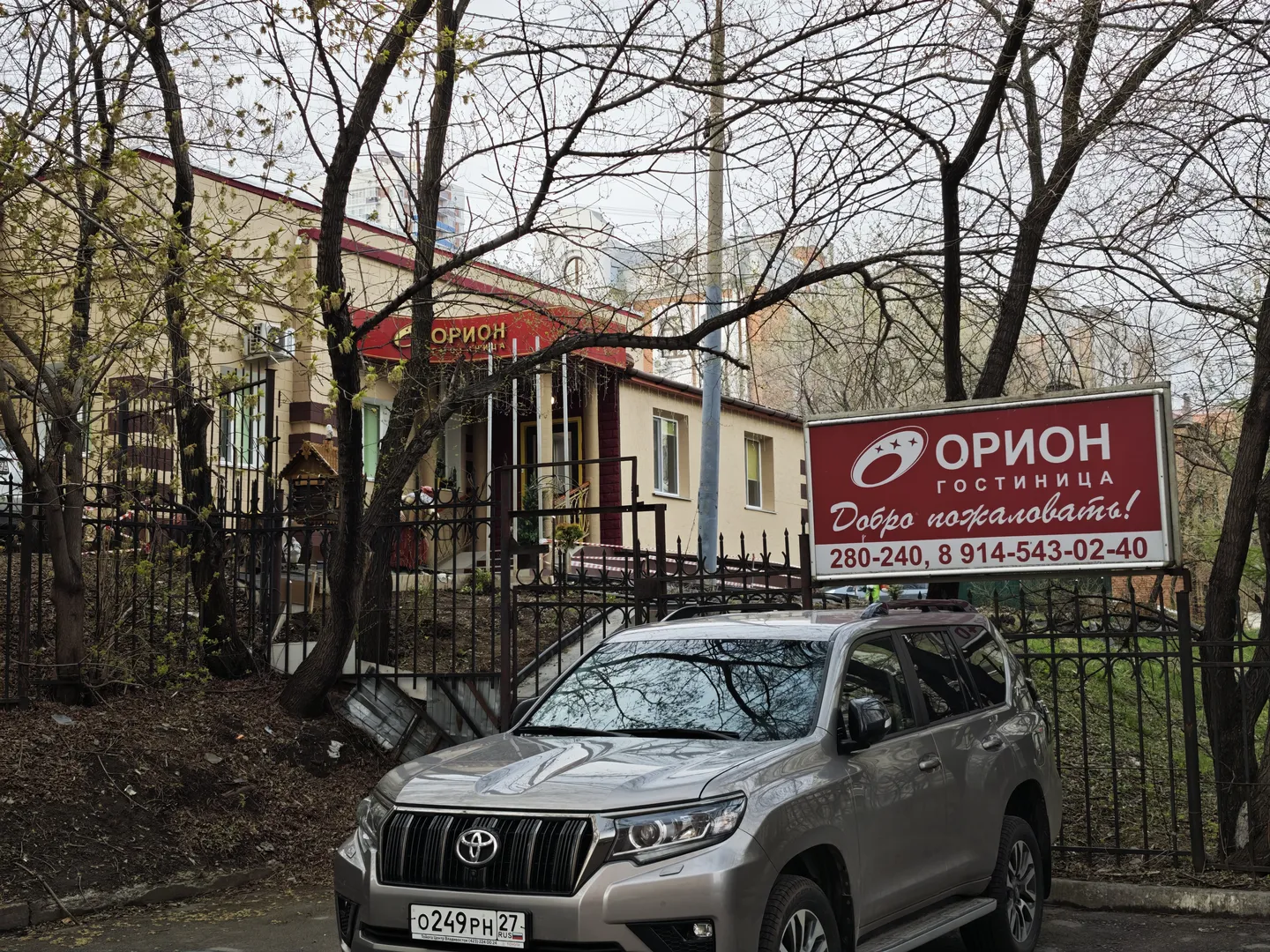 一辆银色SUV停在铁栅栏前,栅栏内是一座建筑物。建筑物上方有红色招牌,上面写着“ОРИОН”。周围有树木,地面覆盖落叶。