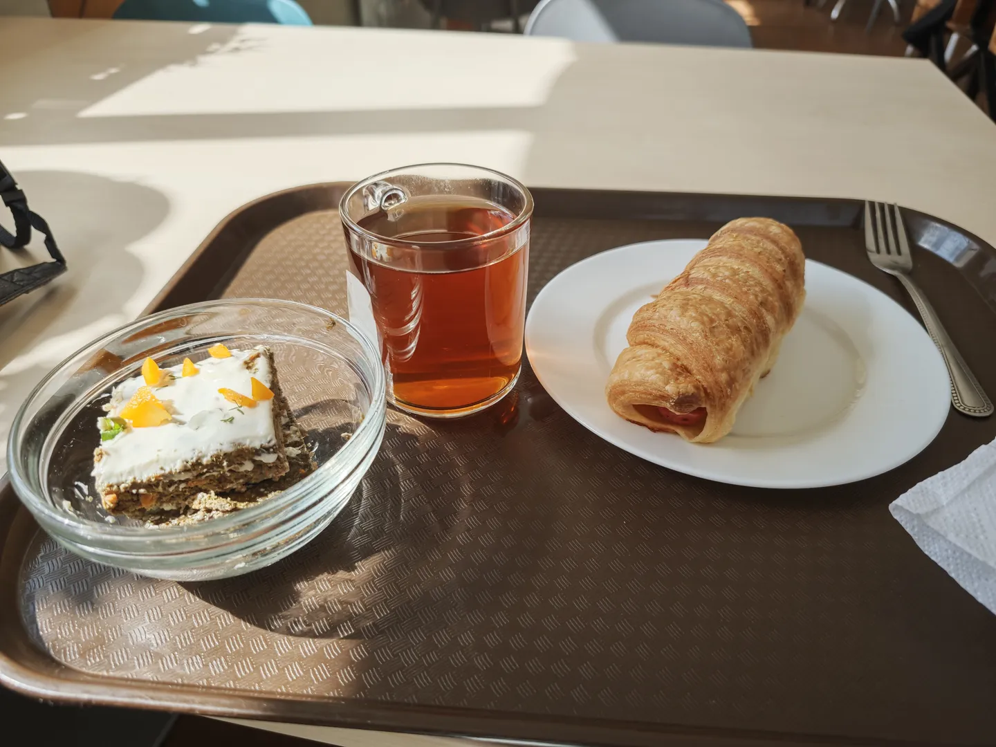 一个托盘上放着一杯茶、一个羊角面包和一块蛋糕。茶杯透明,羊角面包金黄,蛋糕上有奶油和橙片装饰。托盘为棕色,放在白色桌子上。