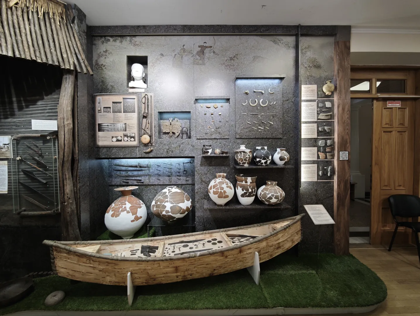 博物馆展示区陈列着古代陶器和工具,墙上挂着各种文物的介绍牌。一个木制独木舟放置在绿色地毯上,旁边是大型陶罐。背景墙上有壁画和雕塑。