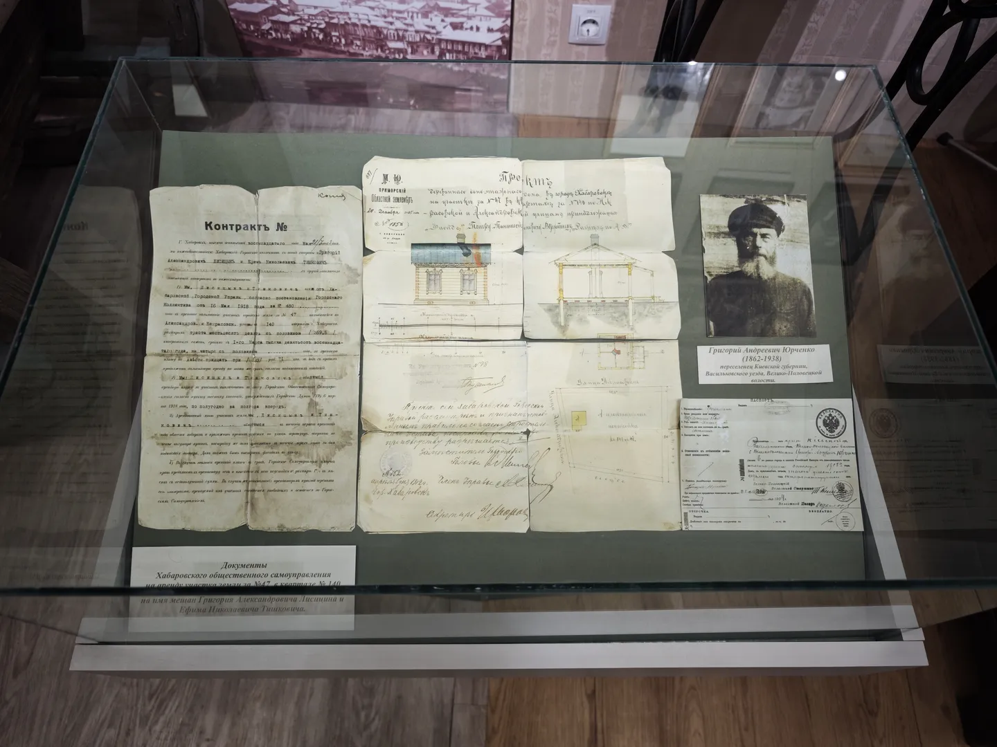 玻璃展柜内展示了几份古老文件和一张人物照片,文件上有手写文字和印章。展柜底部标有说明牌,解释文件的历史背景。背景墙上有建筑物的图片。