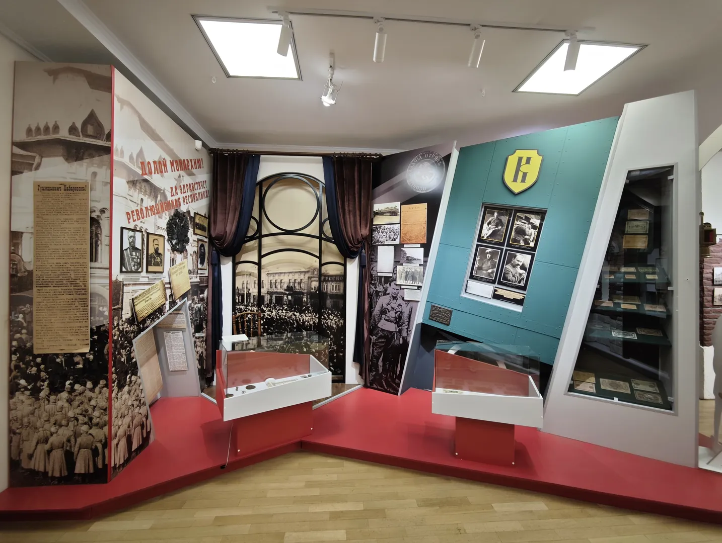 展览区以红色为主色调,墙上挂满了历史照片和标语。展示柜内陈列着各种文件和纪念品,背景墙上装饰有大型历史场景画。整体布局紧凑,信息丰富。