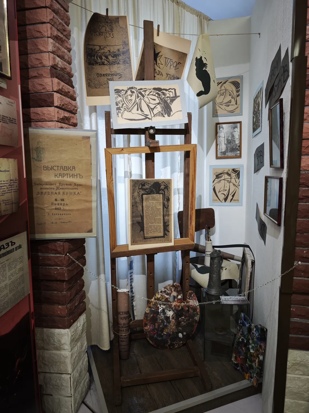 展览区内展示绘画作品和相关物品。墙上挂有多幅画作,前方有画架和调色板。