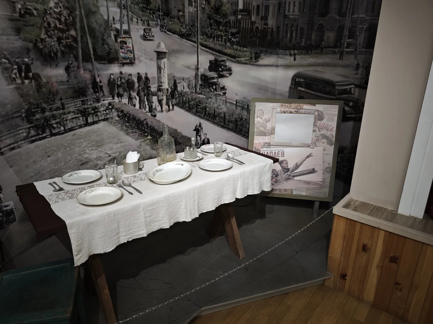 一张桌子铺着白色桌布,上面摆放着盘子、杯子和餐具。背景是一幅黑白城市街景照片,展示了许多行人和车辆。旁边有一个展示板,上面贴有海报和照片。