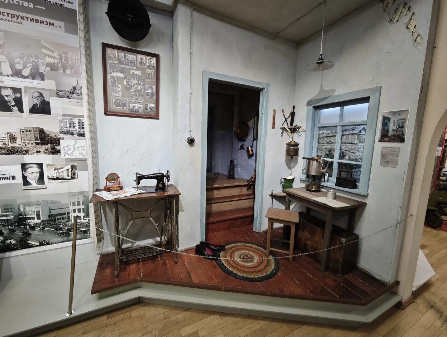 一个小型室内场景模型展示了一个入口区域,左侧有一张桌子,上面放着缝纫机。右侧有一个小桌子,上面放着茶壶和杯子,旁边是一扇窗户。地板上铺着一块圆形地毯。