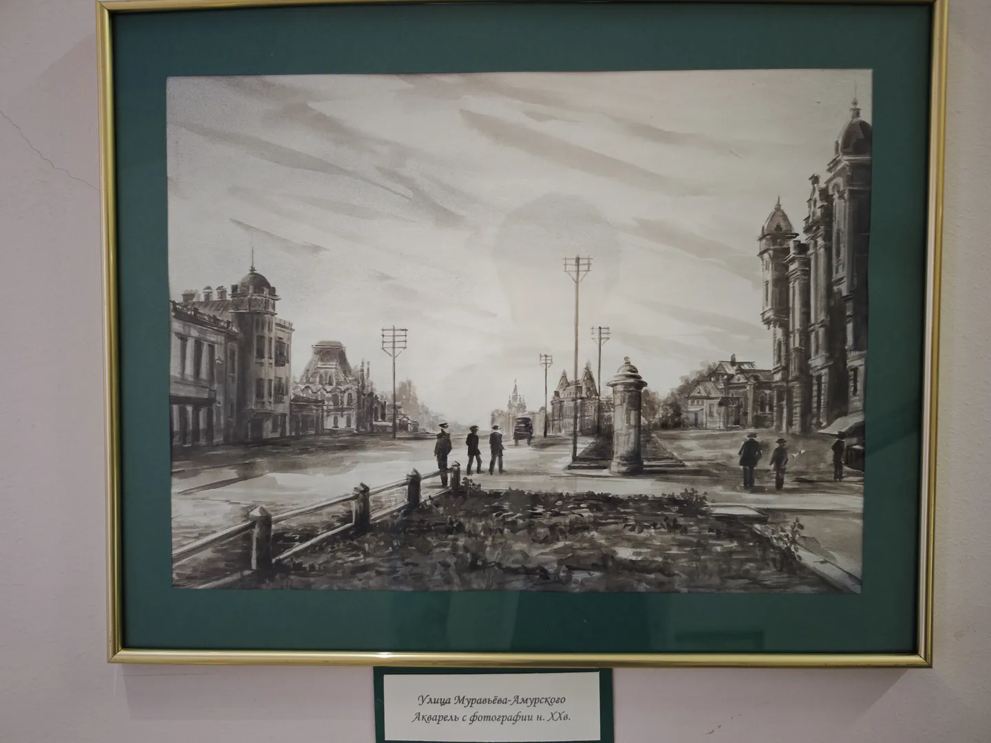 一幅黑白城市风景画挂在墙上,画框为金色。画中描绘了街道、建筑物和行人,天空中有流动的云朵。画下方有一张白色标签,提供画作信息。