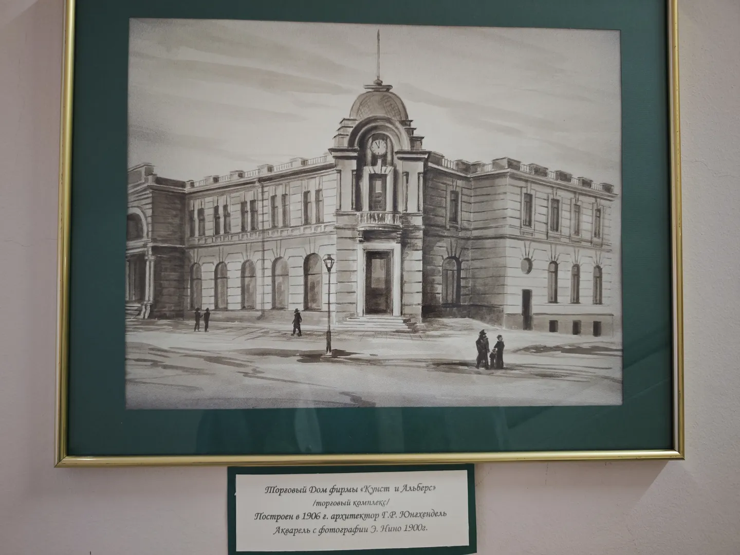 一幅黑白建筑画,展示了一座大型的多层建筑,前面有几个人站立。画作装裱在金色边框内,下方有一段俄文说明文字。