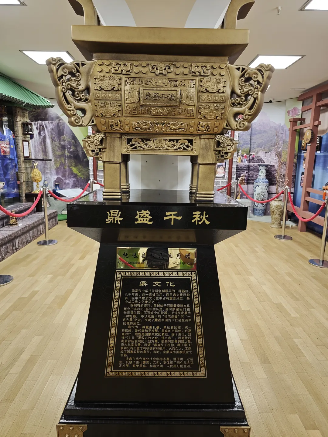 一座大型青铜鼎放置在黑色基座上,基座上有金色文字说明。鼎身雕刻复杂图案,周围有红色绳索围栏。背景为室内展览环境。