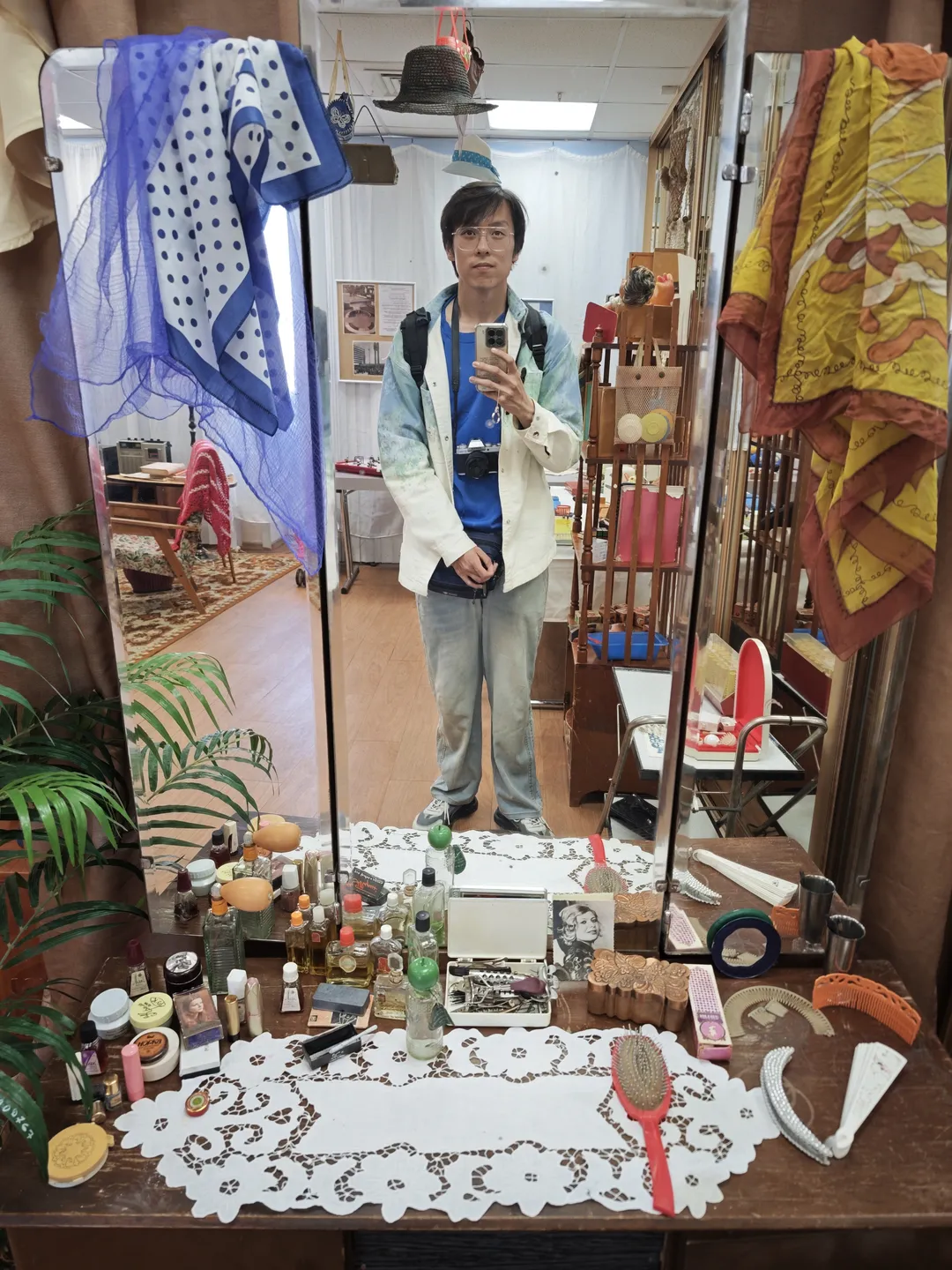 一个人站在镜子前自拍,镜子上方悬挂着几条围巾。镜子下方的桌子上摆放着各种化妆品和梳妆用品。背景中可以看到一些家具和装饰物。
