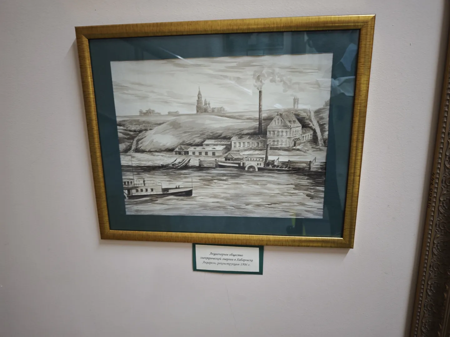 一幅黑白画作描绘了一座河边的工厂建筑,烟囱冒出烟雾,河上有船只。画作被金色边框装裱,下方有一个标签说明。背景为白色墙面。
