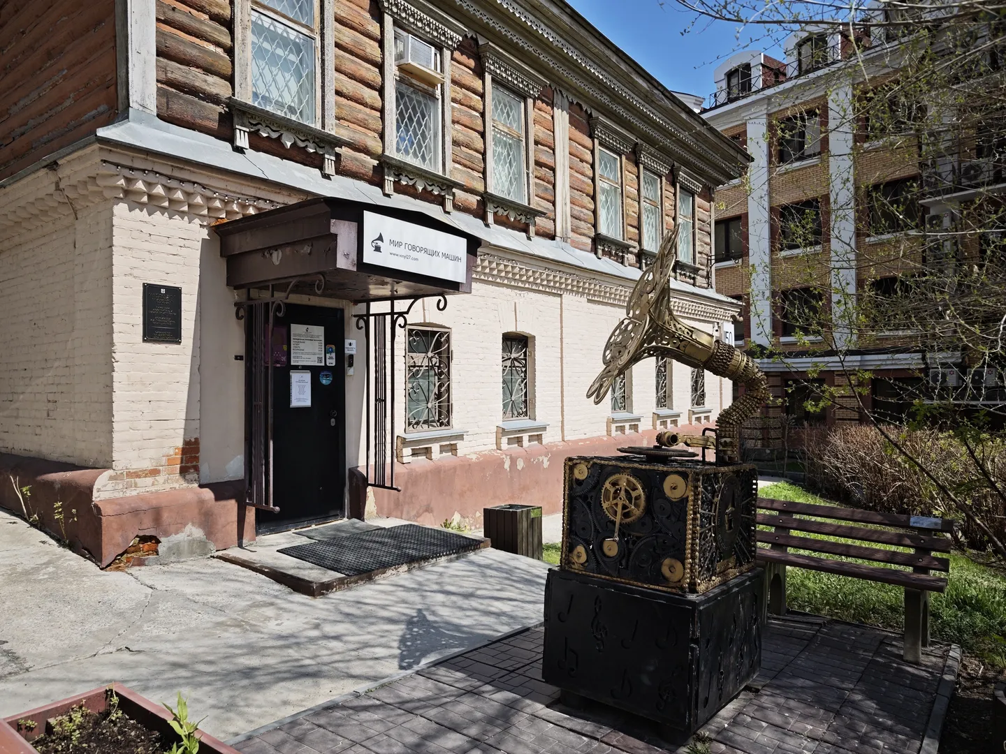 一座木质建筑入口处有一个黑色门,门上方悬挂着标牌。门前有一个金属雕塑,形状像一只展翅的鸟。旁边有一张长椅和一些植物。