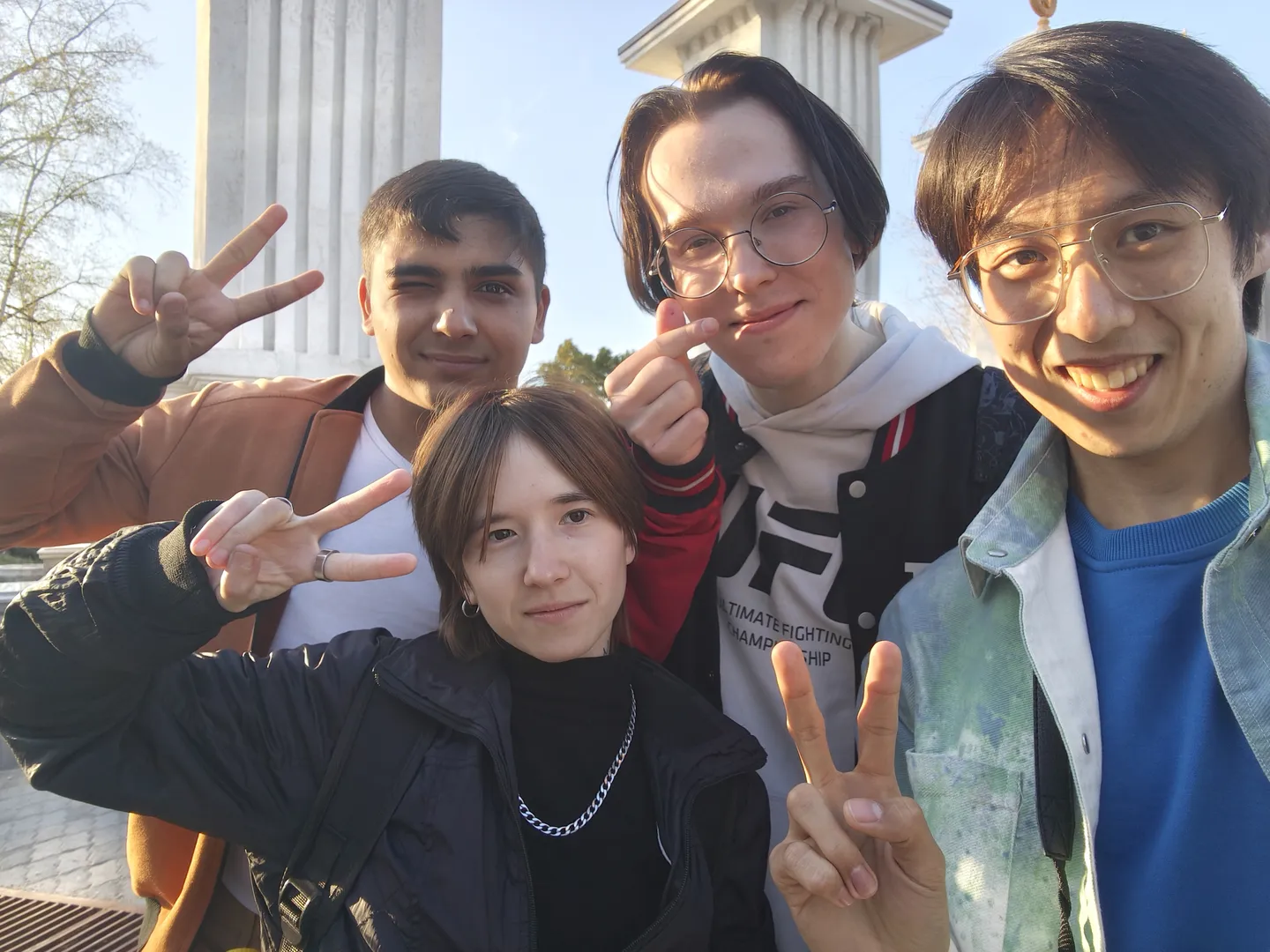 四个人站在一起合影,其中三人比出胜利手势,背景是柱子和天空。