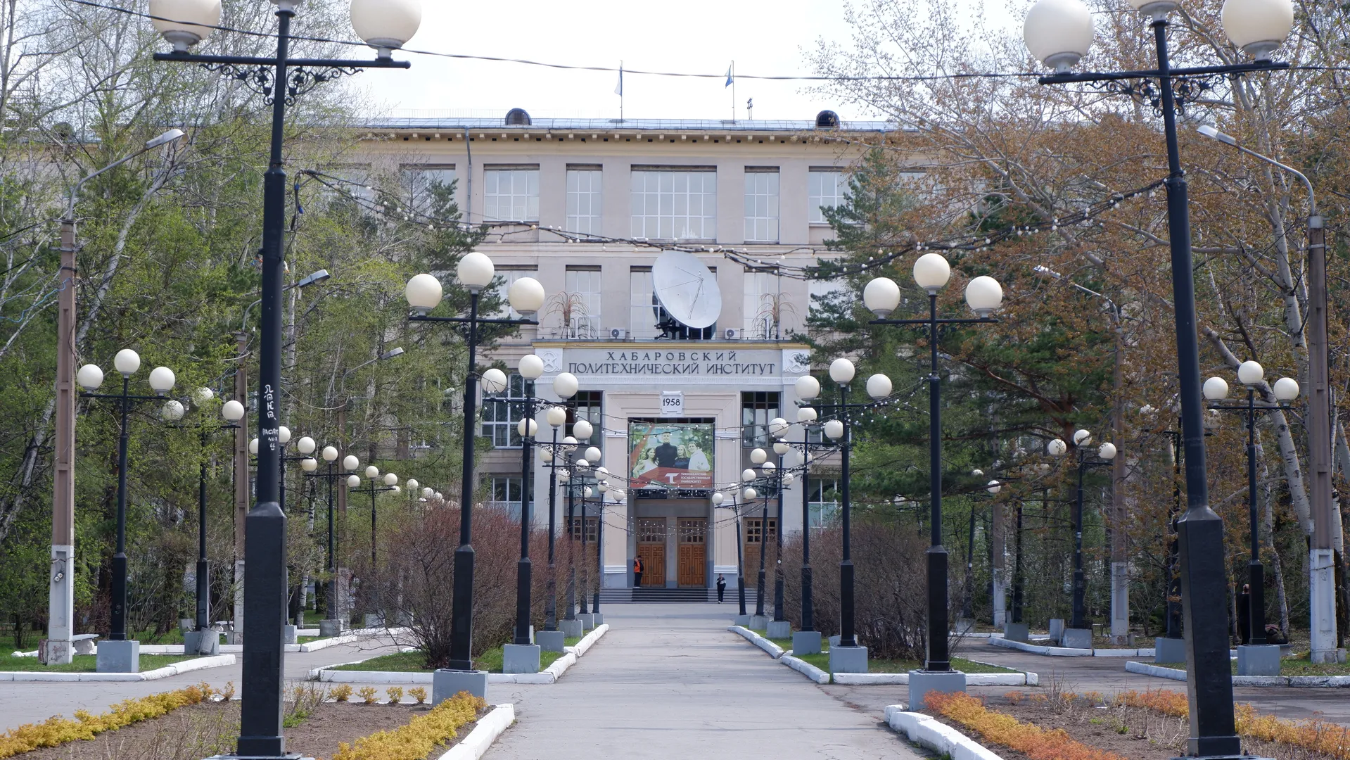 一条铺有路灯的宽阔道路通向一座标有“ХАБАРОВСКИЙ ПОЛИТЕХНИЧЕСКИЙ ИНСТИТУТ”的建筑,建筑前有一个大型卫星天线。道路两旁种植着树木和灌木丛。建筑物的入口处有几扇木质大门。