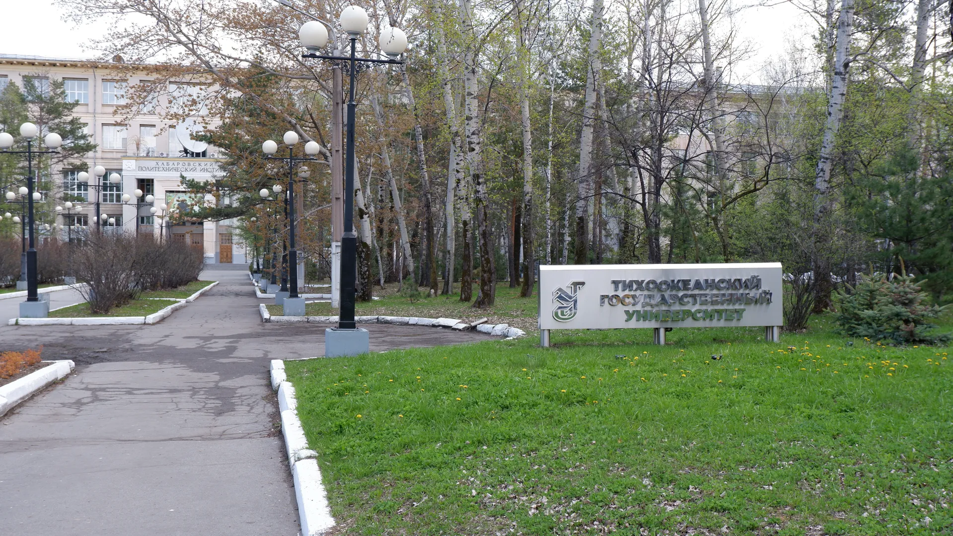 一条铺有路灯的道路旁边有一块白色标志牌,上面写着“ТИХООКЕАНСКИЙ ГОСУДАРСТВЕННЫЙ УНИВЕРСИТЕТ”。道路两旁种植着树木和灌木丛。标志牌左侧有一个图案标志。