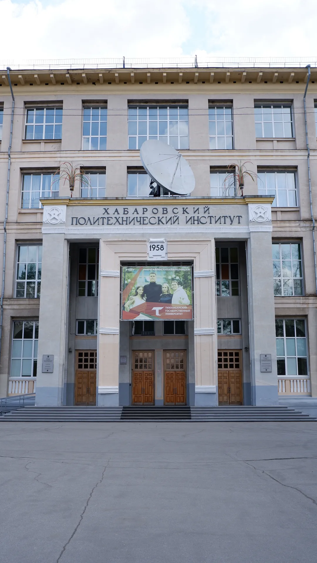 一座标有“ХАБАРОВСКИЙ ПОЛИТЕХНИЧЕСКИЙ ИНСТИТУТ”的建筑,建筑前有一个大型卫星天线。建筑入口处有几扇木质大门。大门上方挂着一幅海报,显示几个人的形象。