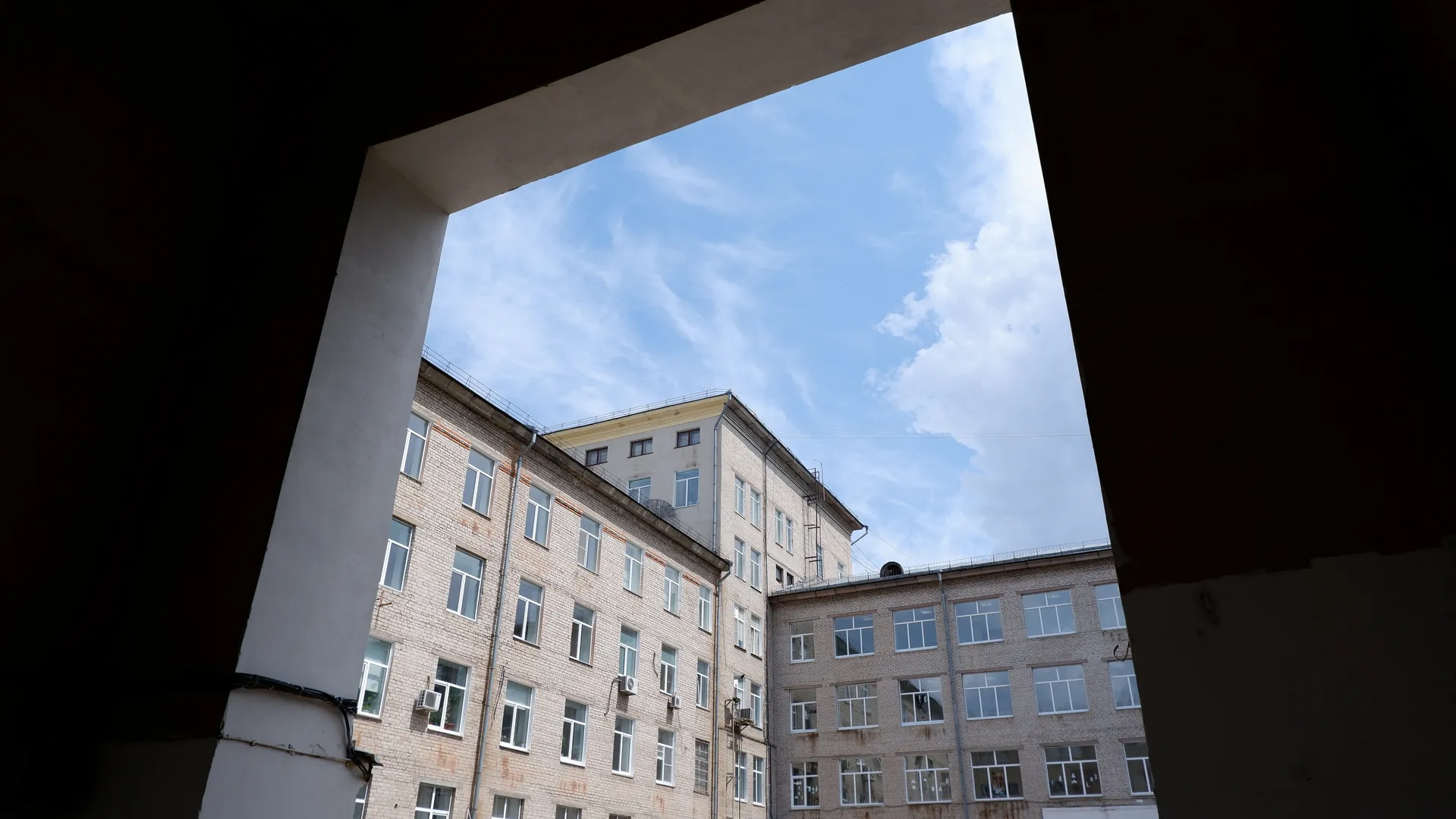 从一个门洞望出去,可以看到一座多层砖砌建筑,建筑上有许多窗户。建筑侧面有管道和空调外机。天空中有云彩。