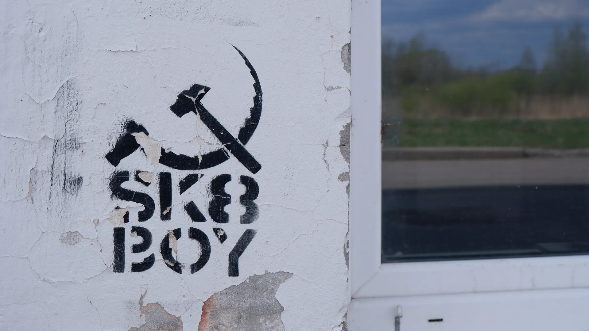 墙上有一个黑色涂鸦,图案为镰刀锤子标志,下方写着“SK8 BOY”。墙面有剥落的痕迹,显示出一定的磨损。背景中可以看到一片绿色的草地和树木。