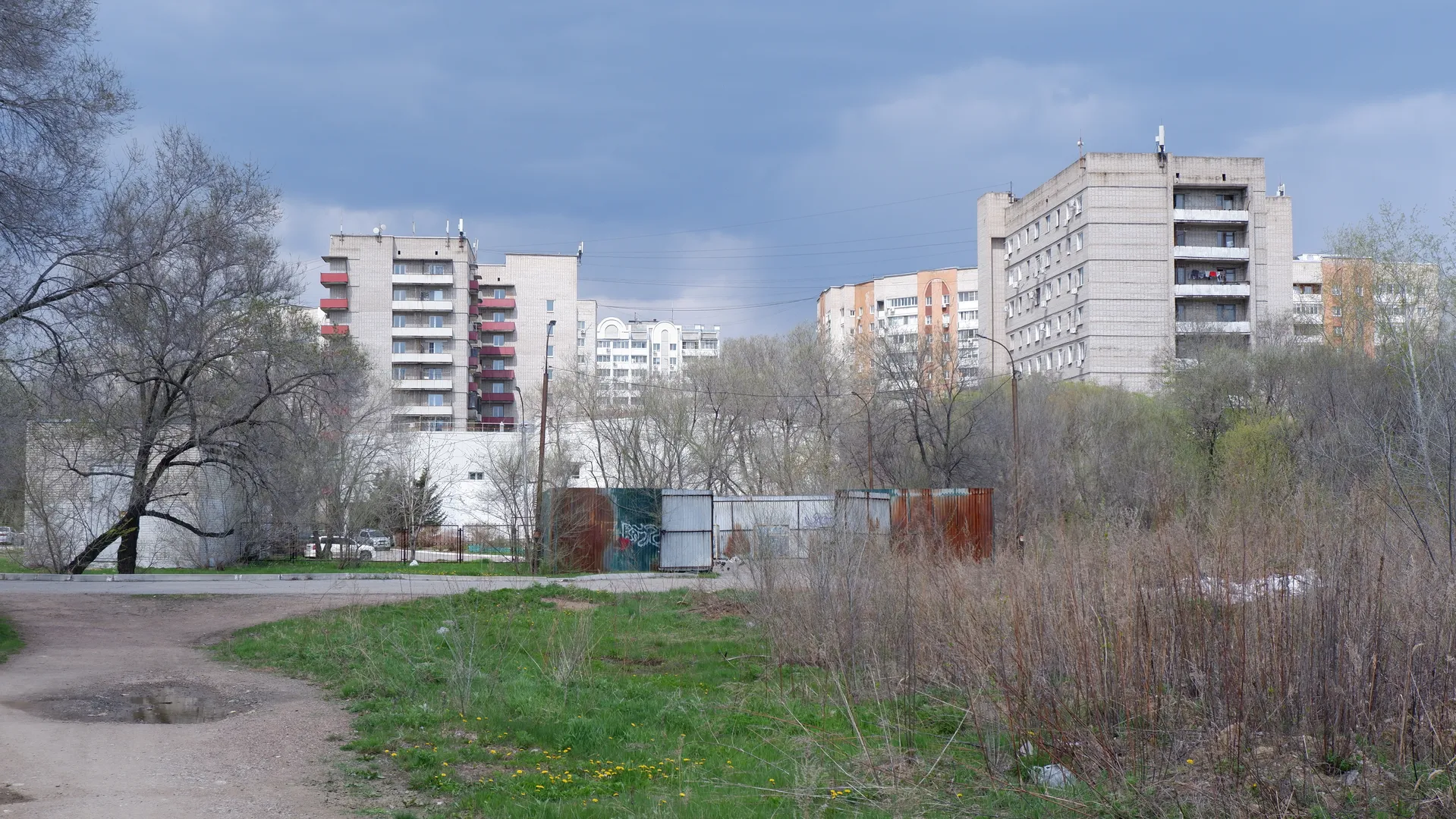 多层建筑群后方是树木和灌木丛,前景是一片草地。建筑物外墙颜色多样,窗户排列整齐。天空中有云层,但光线充足。
