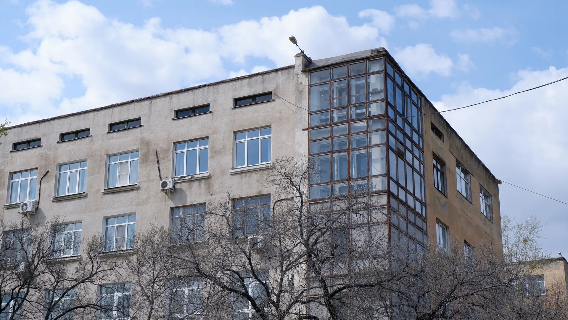 一栋多层建筑的外墙为浅色,角落处有大面积的玻璃窗。建筑顶部有一个灯饰,窗户排列整齐。前景中有几棵树,树枝光秃。