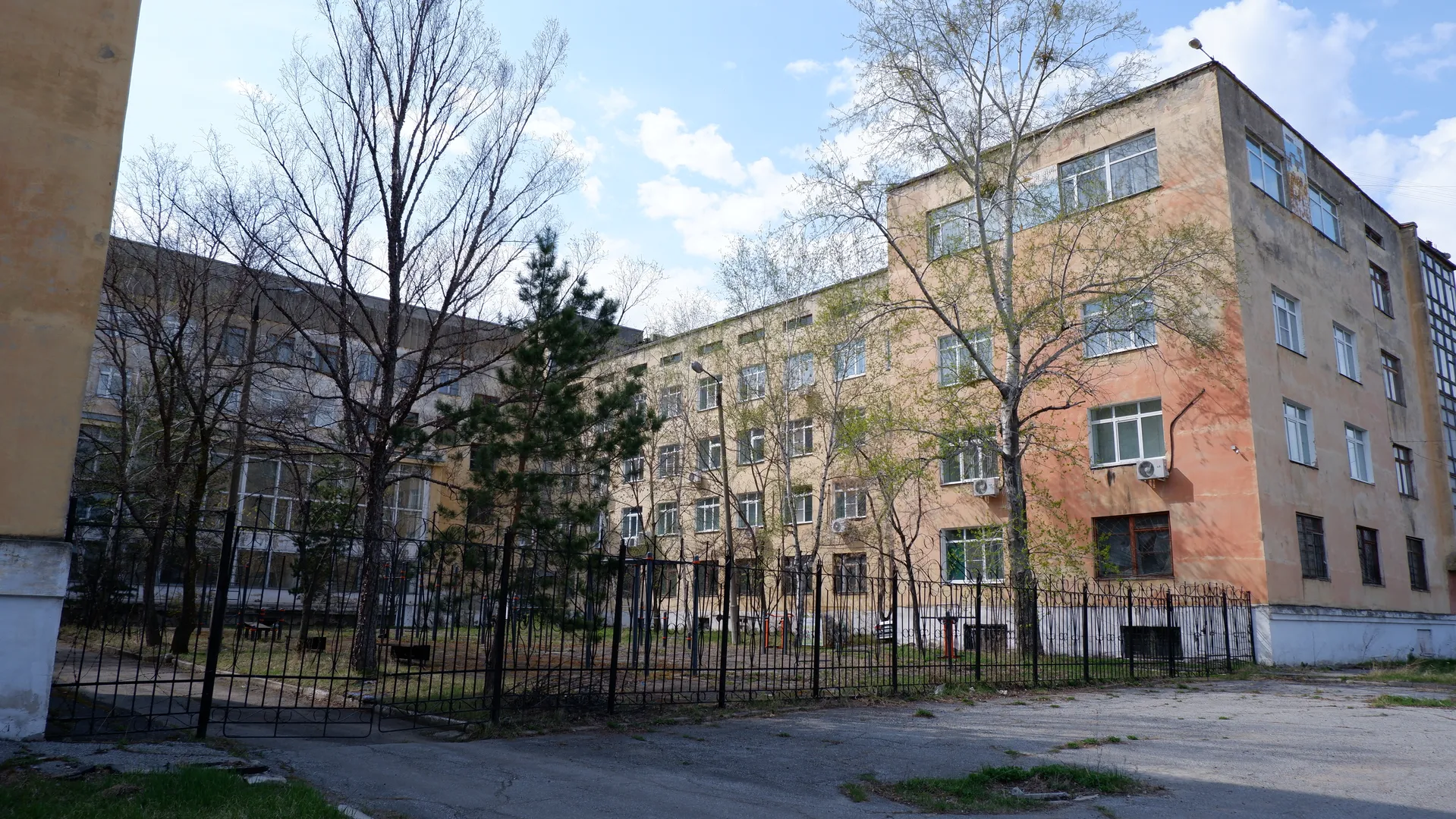 一栋多层建筑前有铁栅栏和几棵树。建筑外墙为浅色,窗户排列整齐。前景中有一条小径,地面为水泥材质。