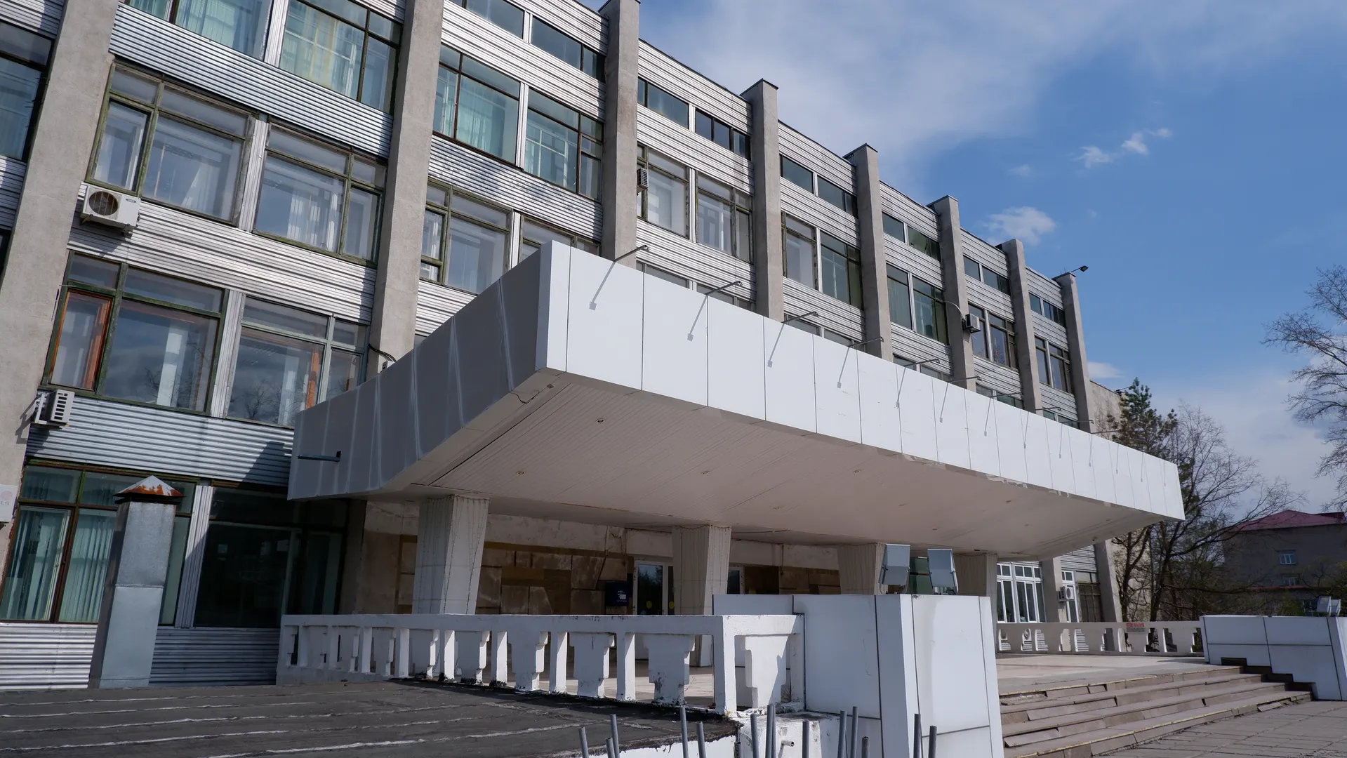一座现代建筑的入口处有一个大型遮阳棚,建筑物有多层窗户和空调设备。入口前方有台阶和护栏,天空晴朗。
