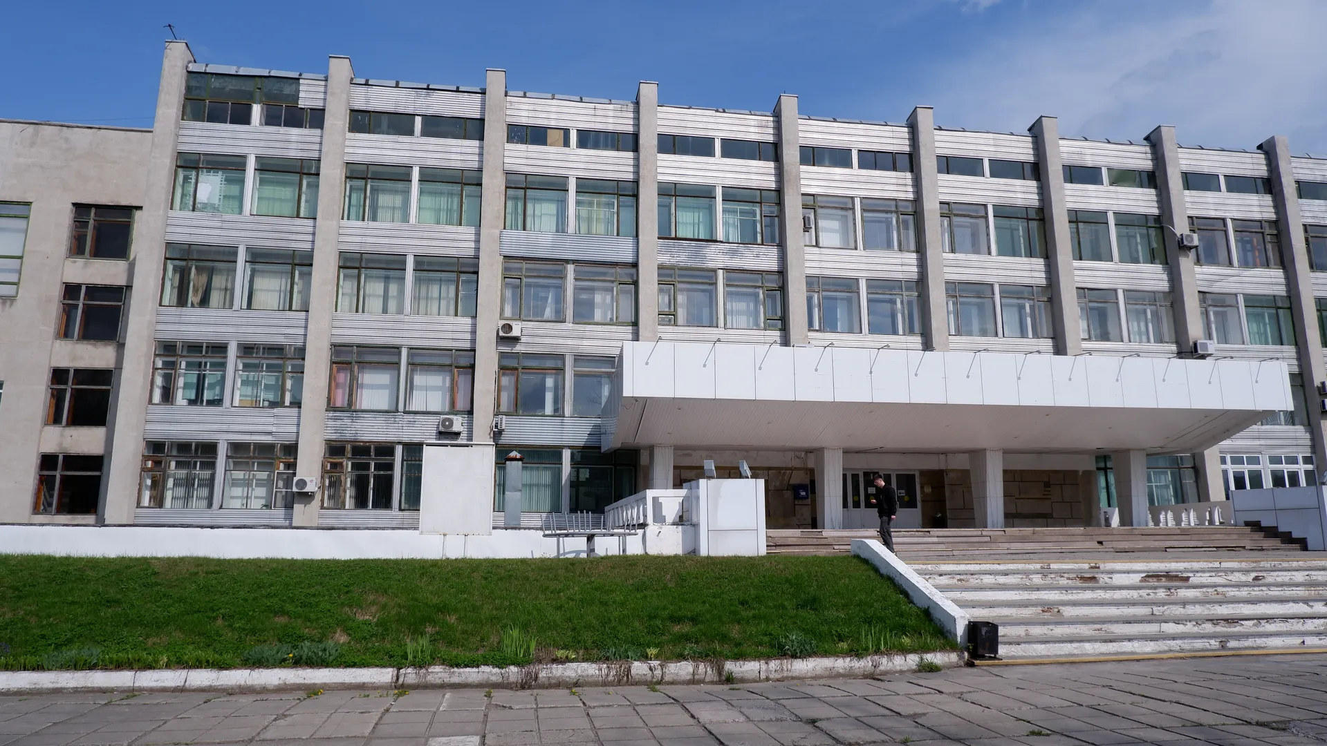 一栋多层建筑正面有许多窗户,外墙为浅色。建筑前有台阶和护栏,一个人站在台阶上。天空晴朗。