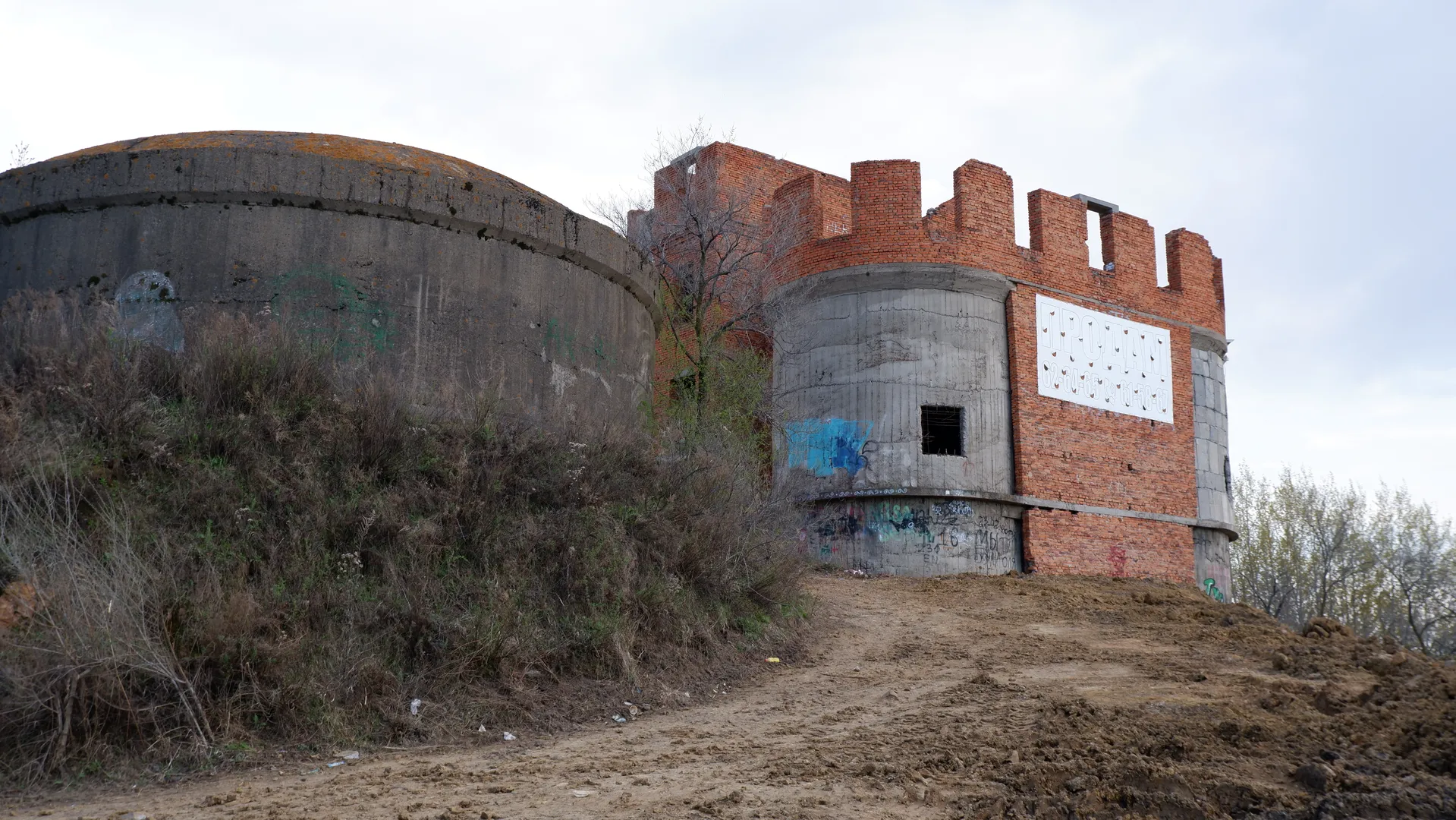 一座未完工的砖砌建筑位于土坡上,部分结构由混凝土构成。建筑上有涂鸦,周围长有杂草。天空阴沉。