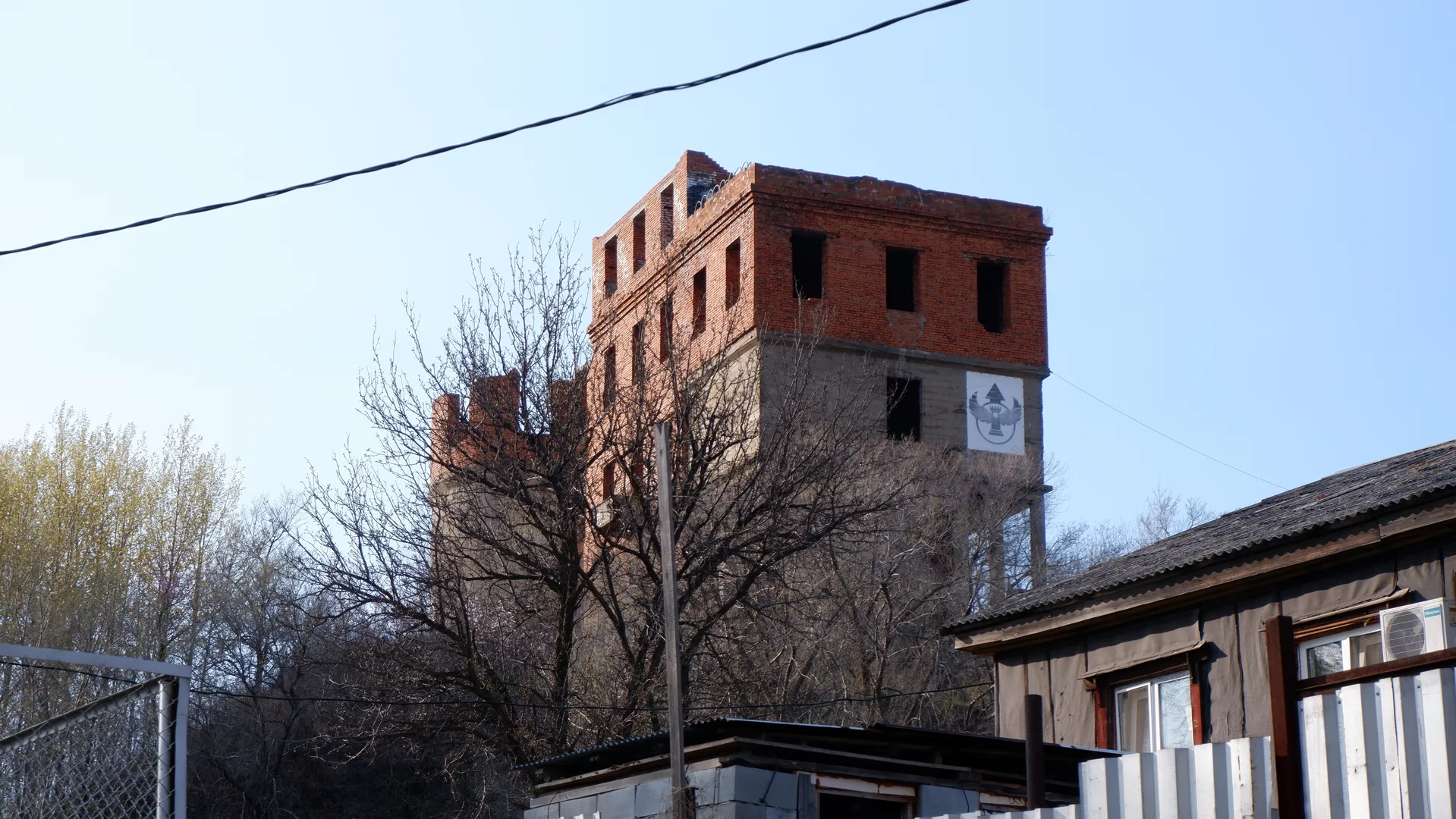 一座高大的红砖建筑顶部残缺,周围有光秃秃的树木。建筑侧面有一个白色标志,上面有图案。天空晴朗。