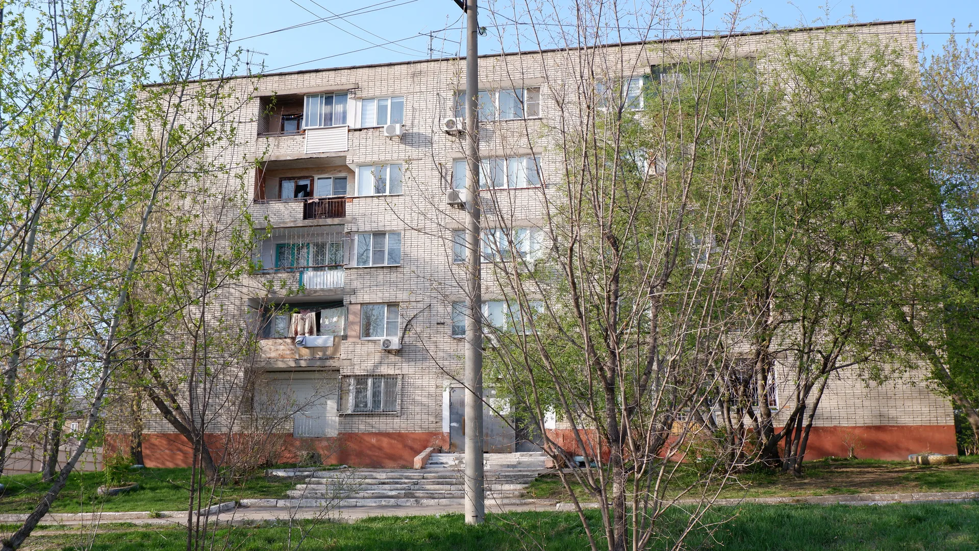一栋多层住宅楼,外墙为浅色砖块,窗户排列整齐。楼前有几棵树,树叶茂盛,台阶通向入口。电线杆和电线穿过画面。