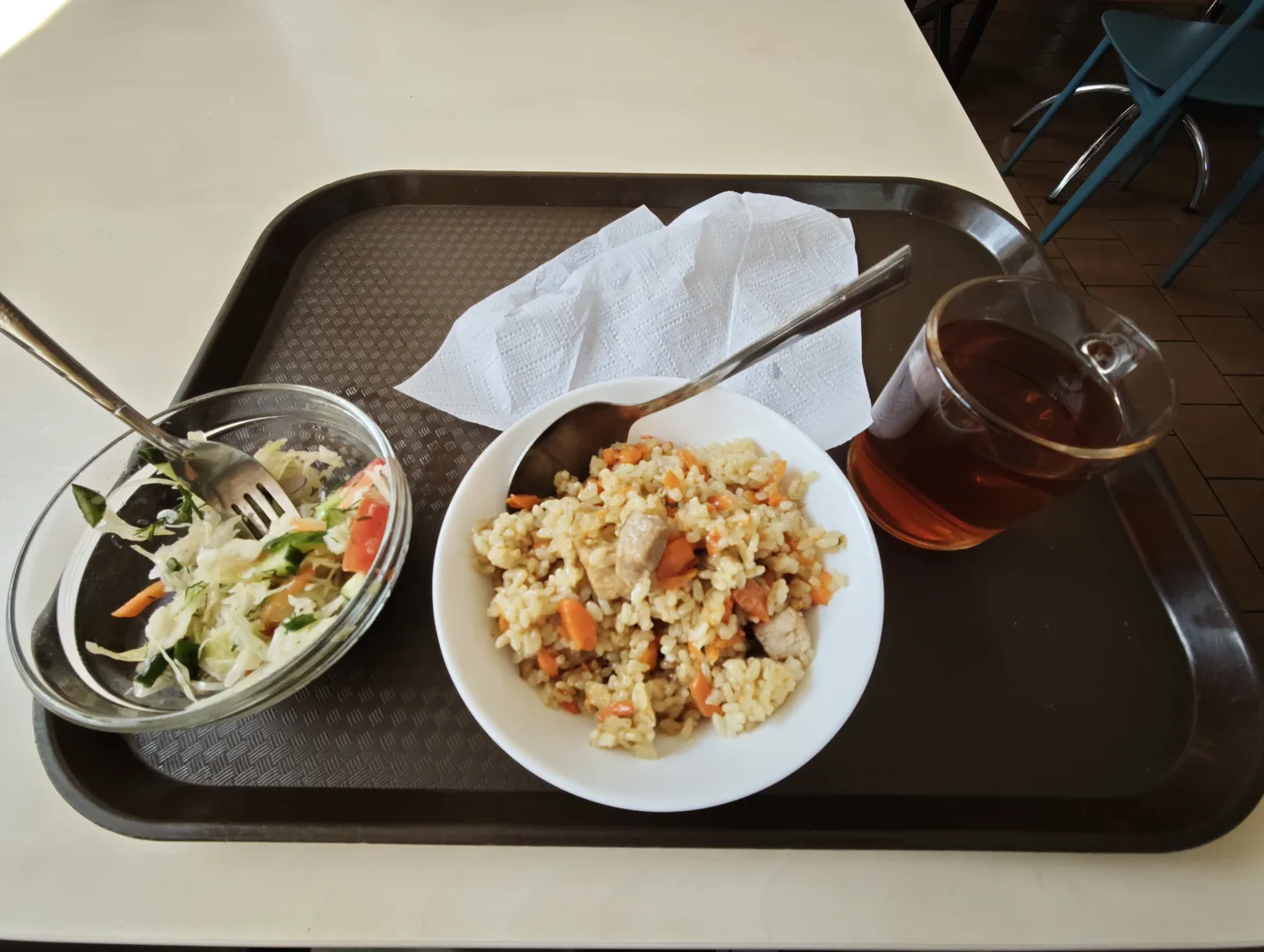 托盘上有一碗米饭、一碗沙拉和一杯茶,旁边放着纸巾和餐具。米饭中有胡萝卜和肉块,沙拉包含蔬菜。托盘放置在桌子上。